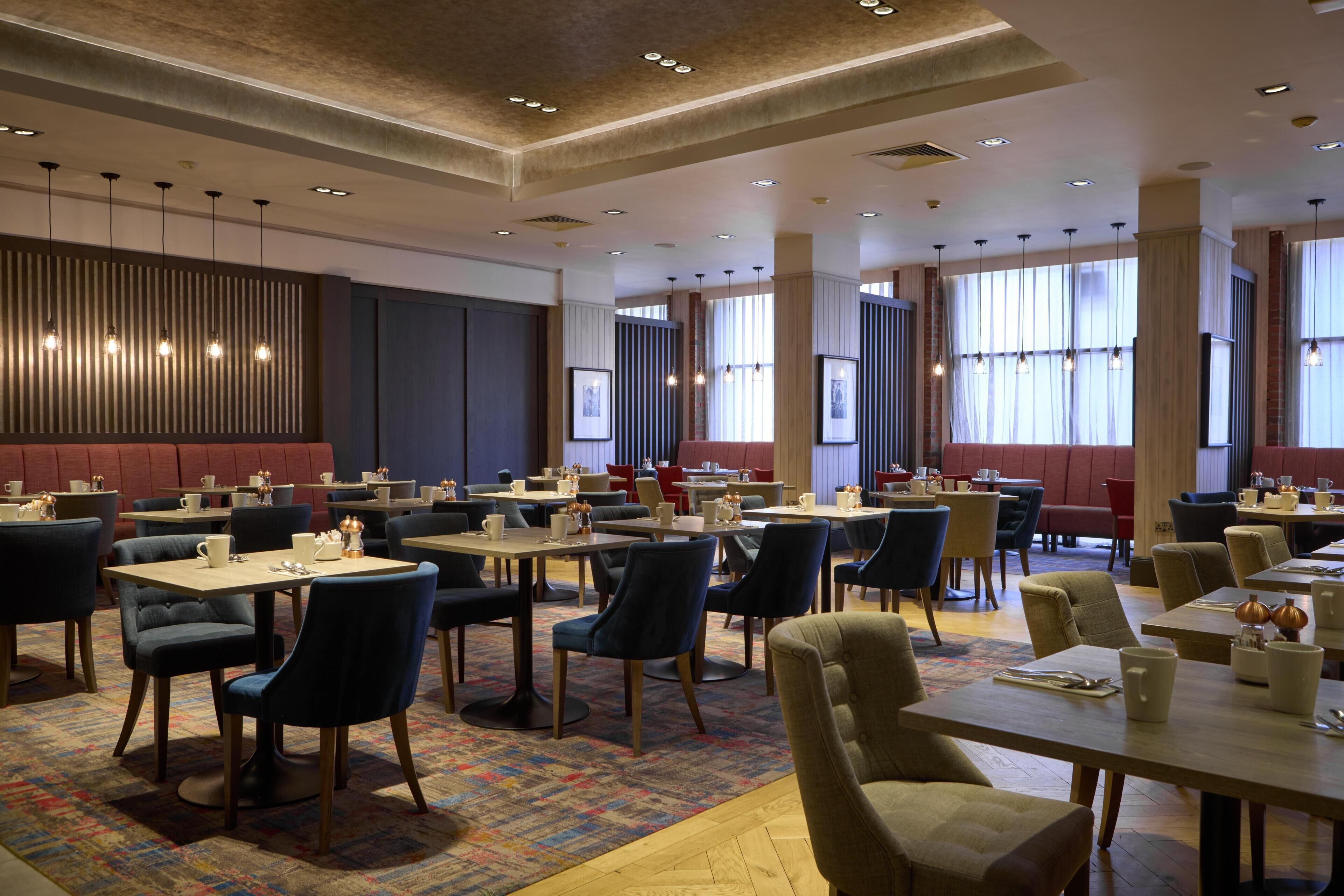 A hotel dining area with multiple tables and chairs, large windows, and a patterned carpet.