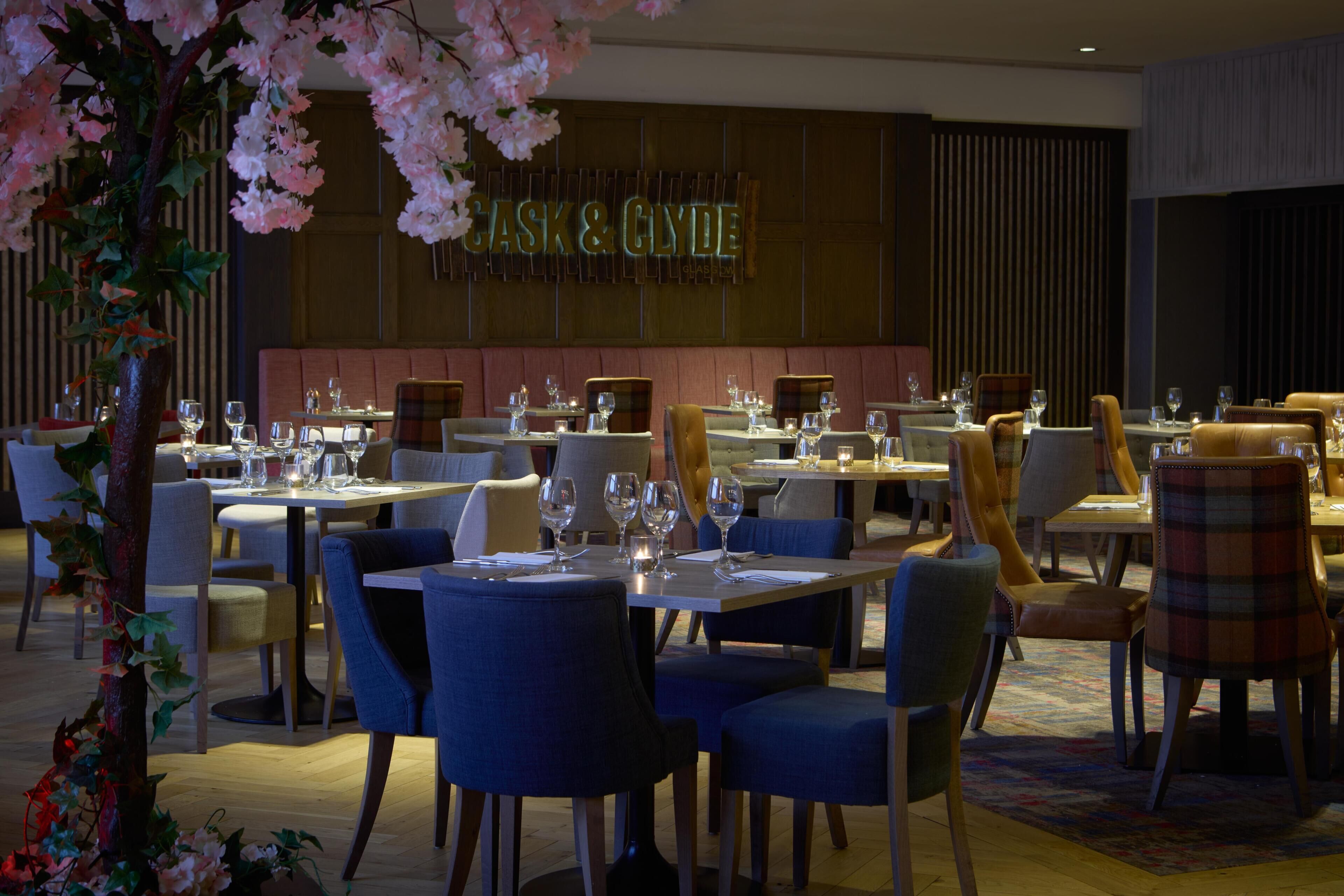 A restaurant interior with tables set for dining, a sign reading 'CASK & CLYDE,' and a decorative tree with pink flowers.