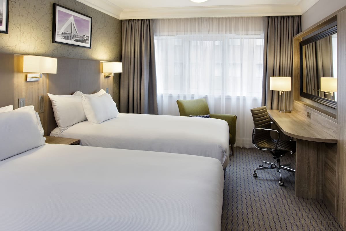 Een hotelkamer met twee bedden, een bureau, een stoel en een groot raam met gordijnen.