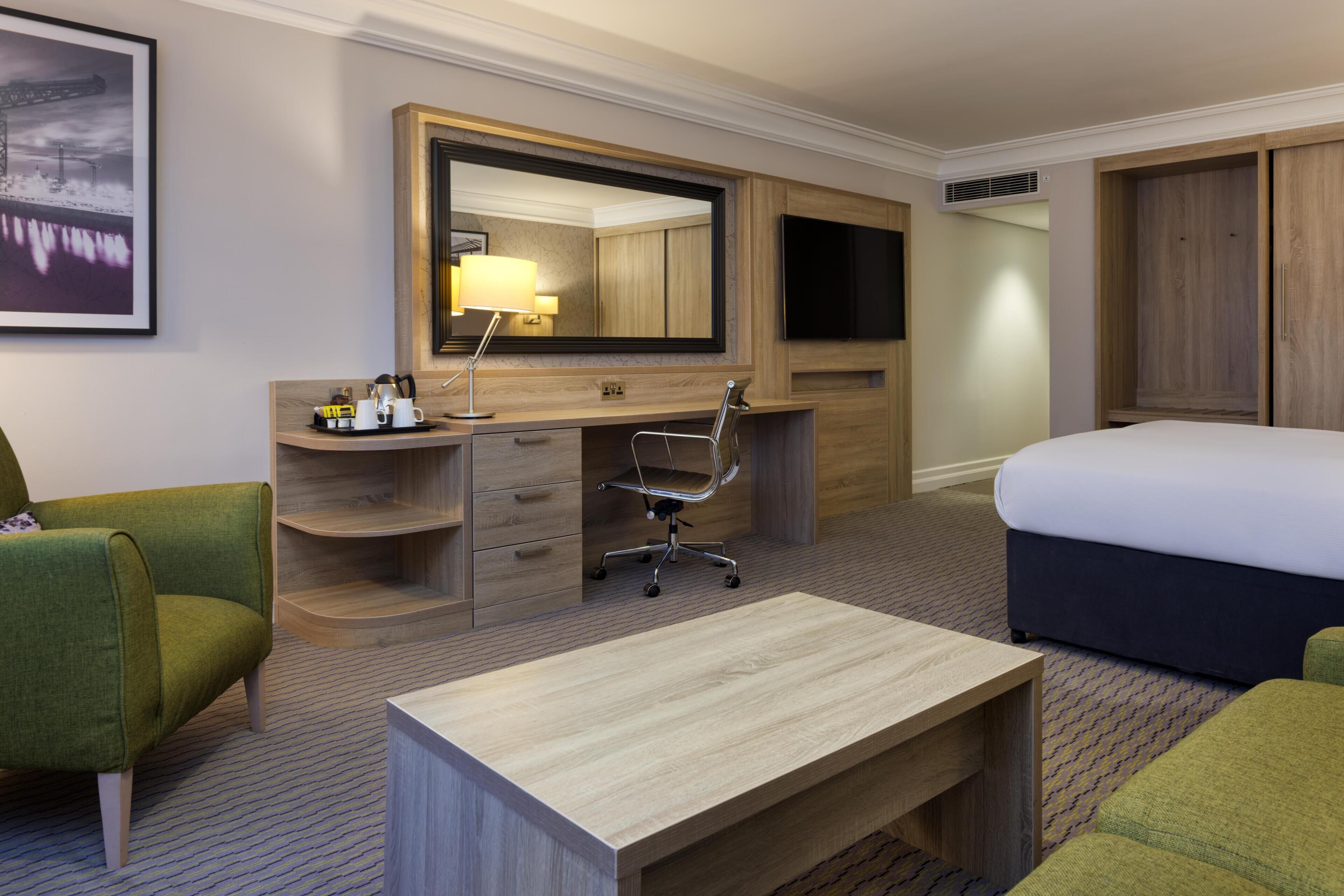 A hotel room with a bed, a desk, a chair, a large mirror, a TV, and a wardrobe.