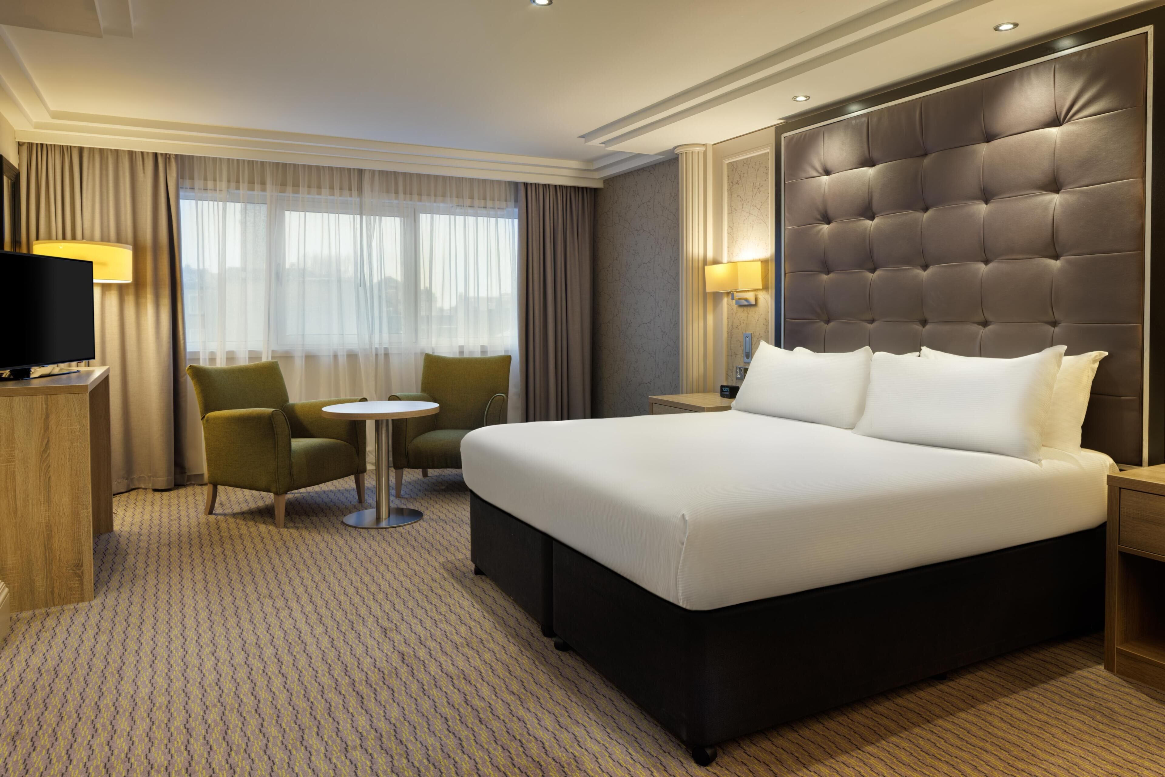 A hotel room with a large bed, a television, a small round table, a chair, and a sofa.