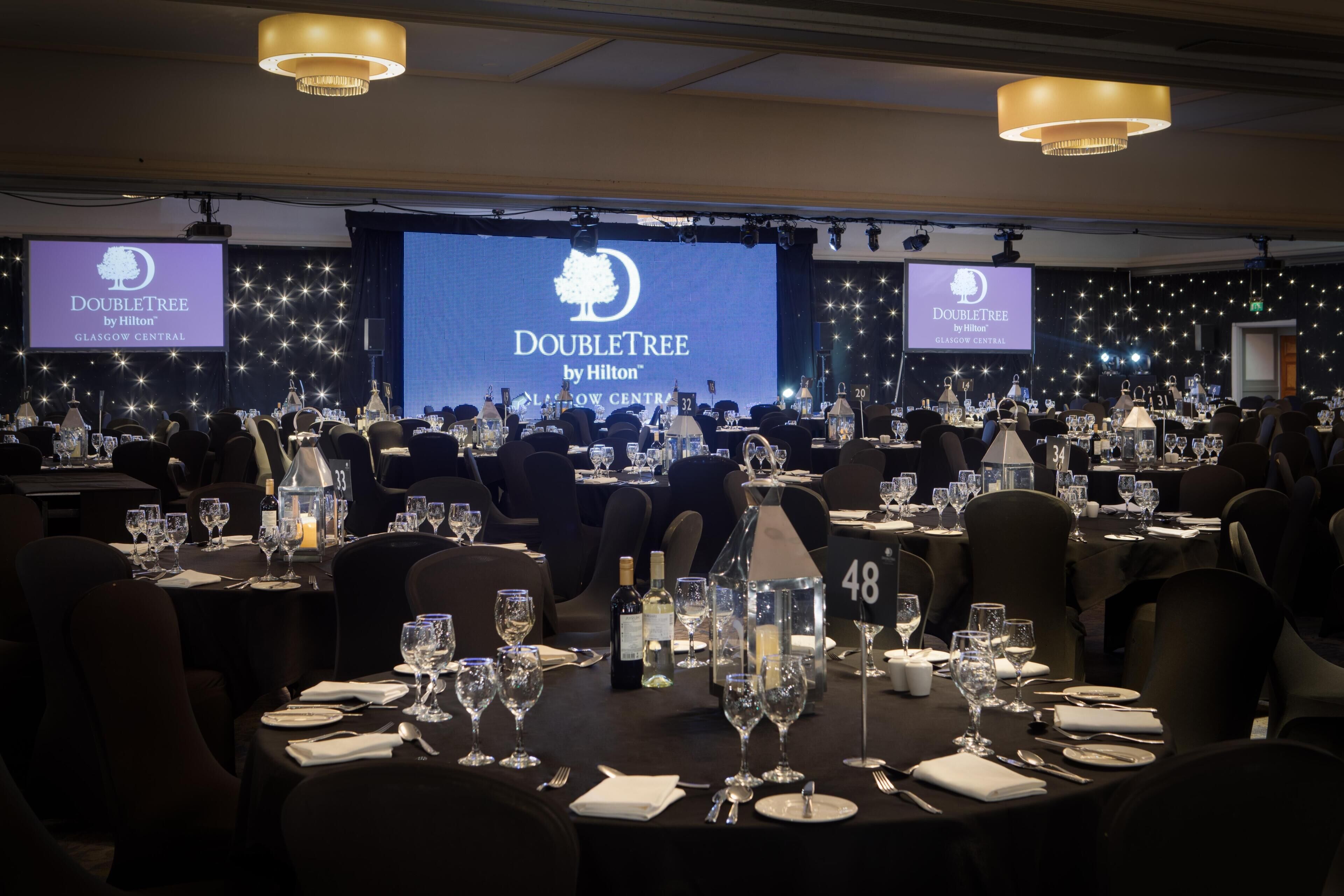 A banquet hall with tables set for a formal event, a large screen displaying the event name, and a chandelier overhead.