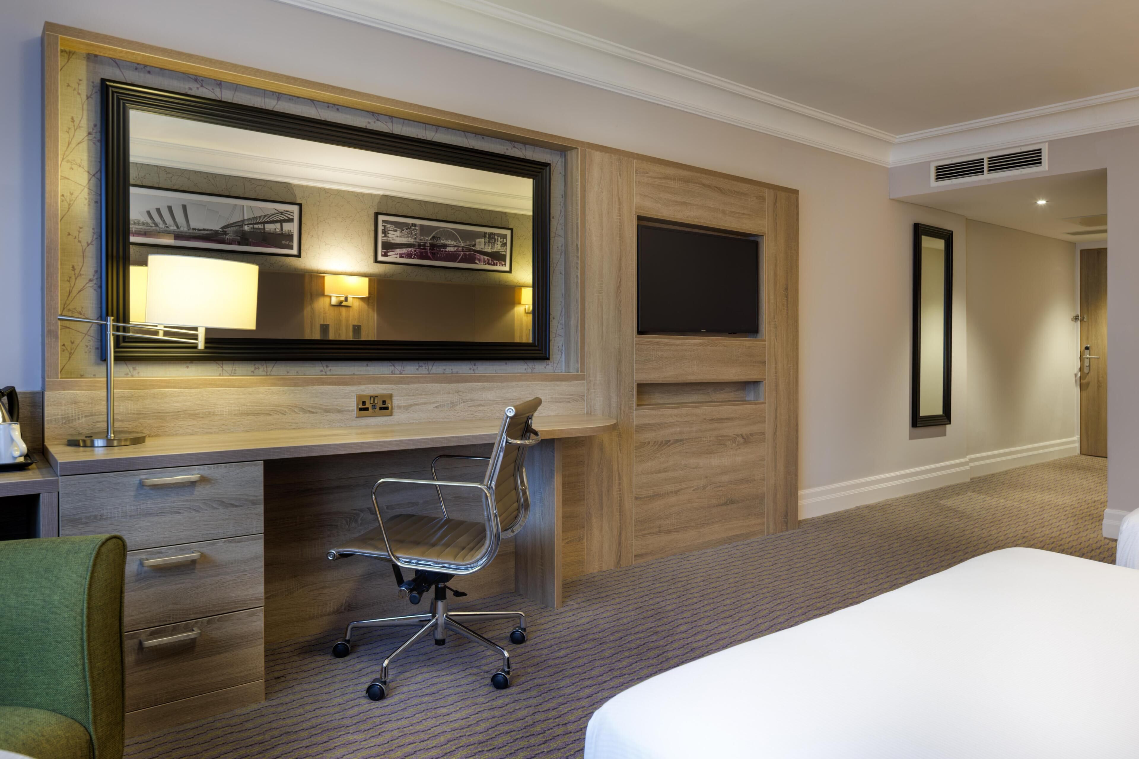 A hotel room with a large bed, a desk with a chair, a large mirror, and a television.