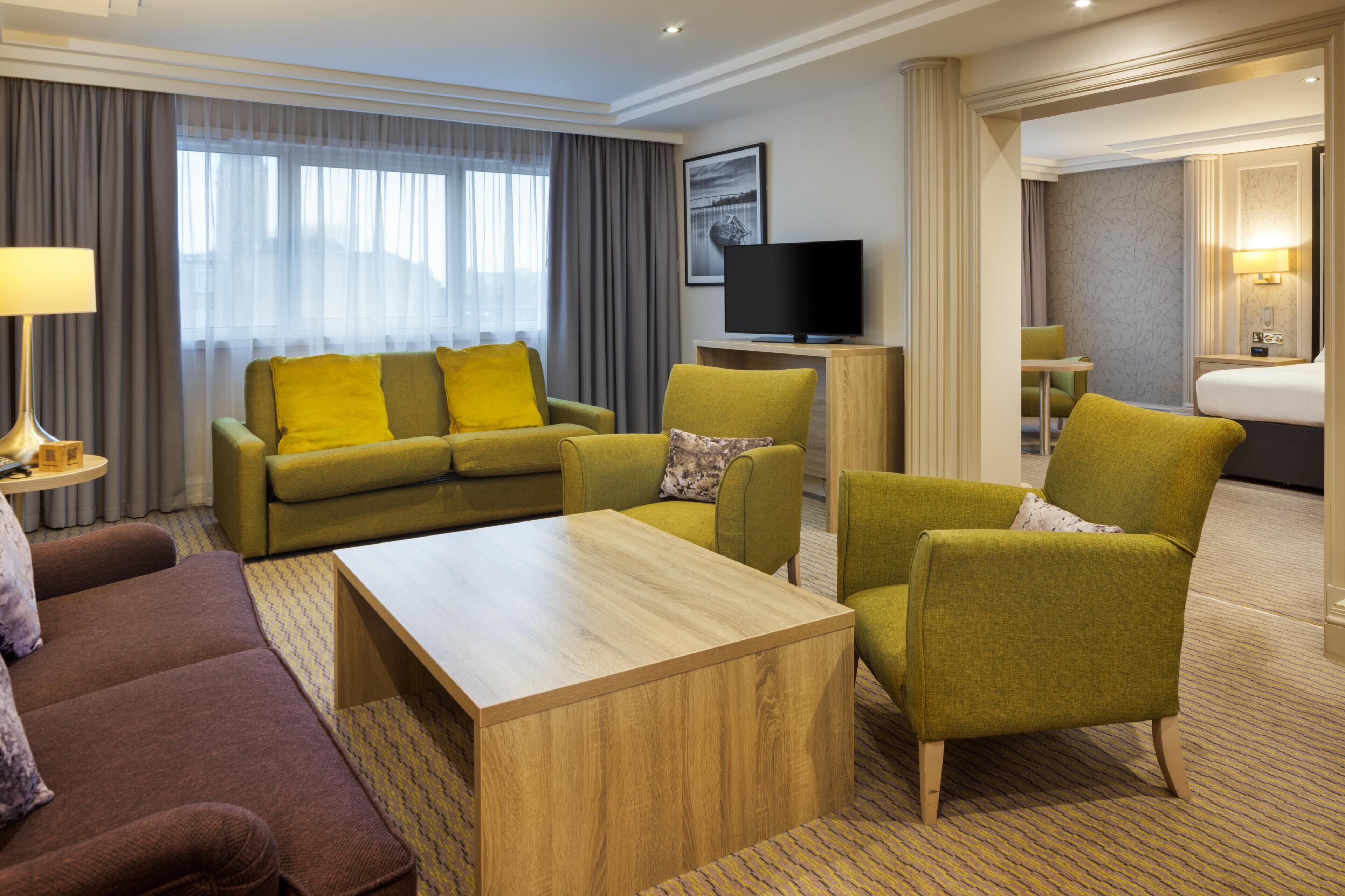 A hotel room with a sofa, armchairs, a coffee table, a TV, a bed, and a bathroom.