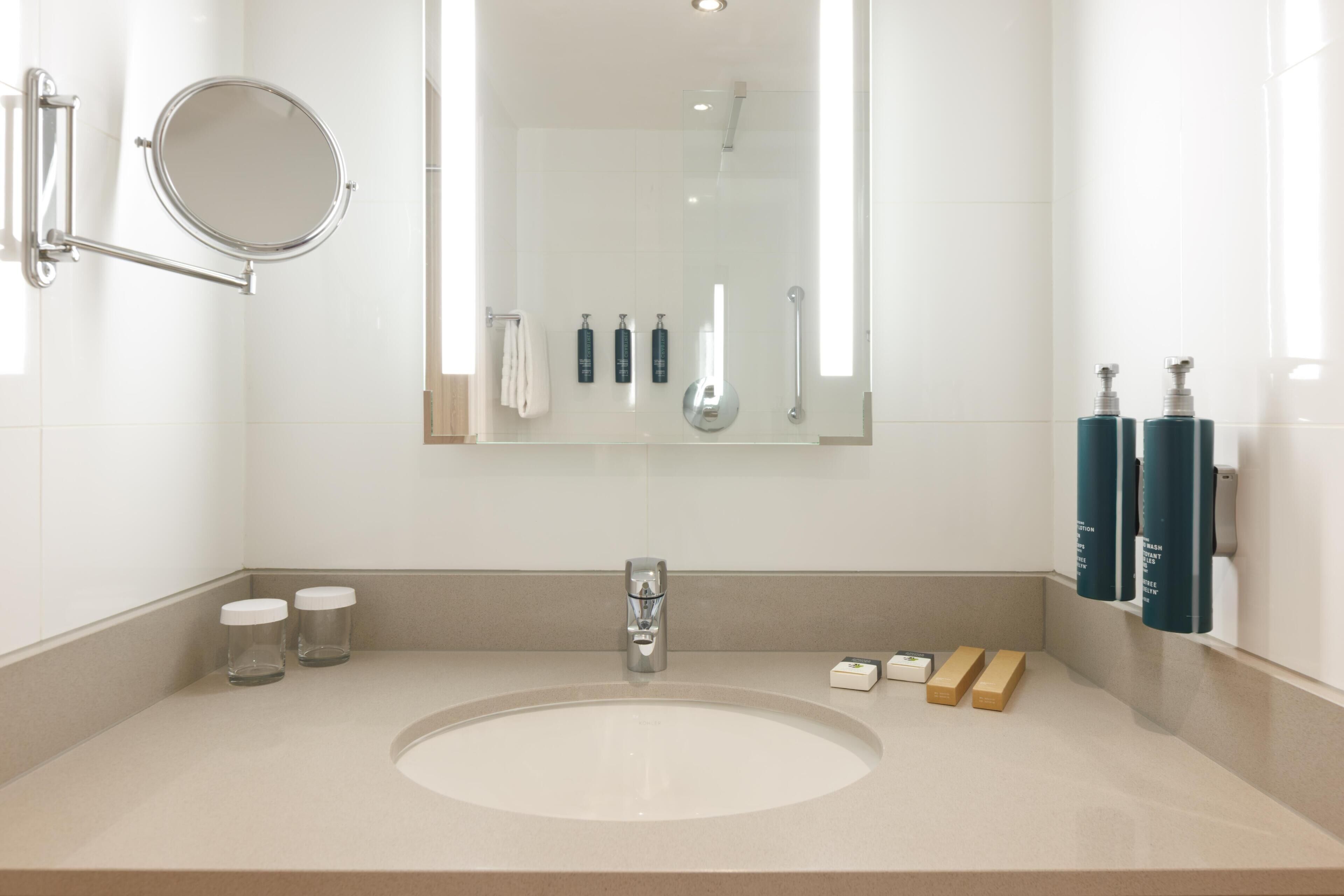 Ein modernes Badezimmer mit einem runden Spiegel, einem Waschbecken, einer Handbrause und Pflegeprodukten an der Wand.