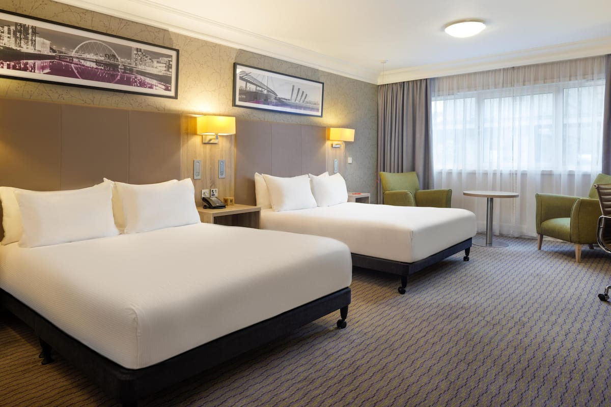 Een hotelkamer met twee bedden, een tafeltje, twee stoelen en een groot raam met gordijnen.