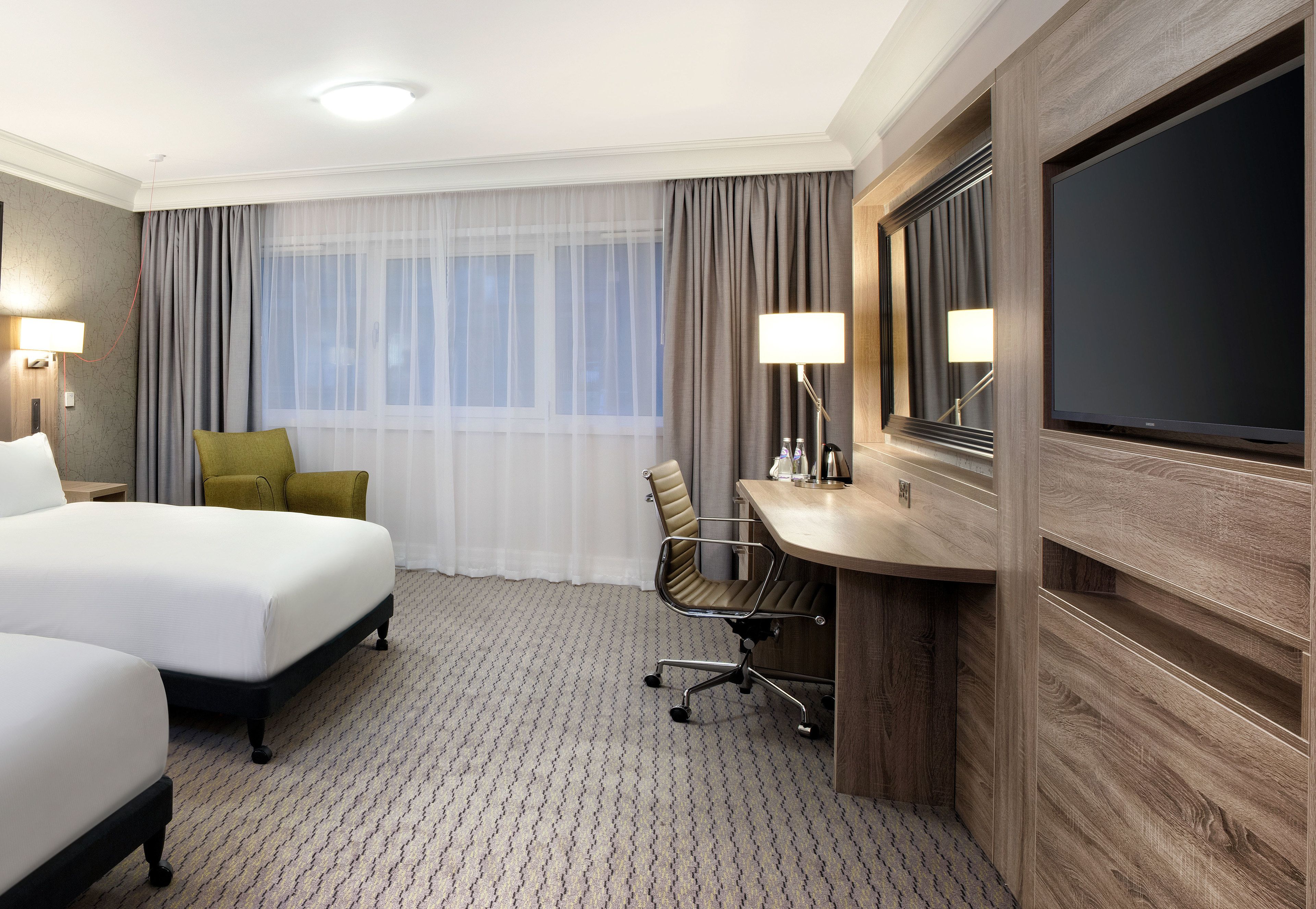 A hotel room with two beds, a desk, a chair, a TV, and a large window with curtains.