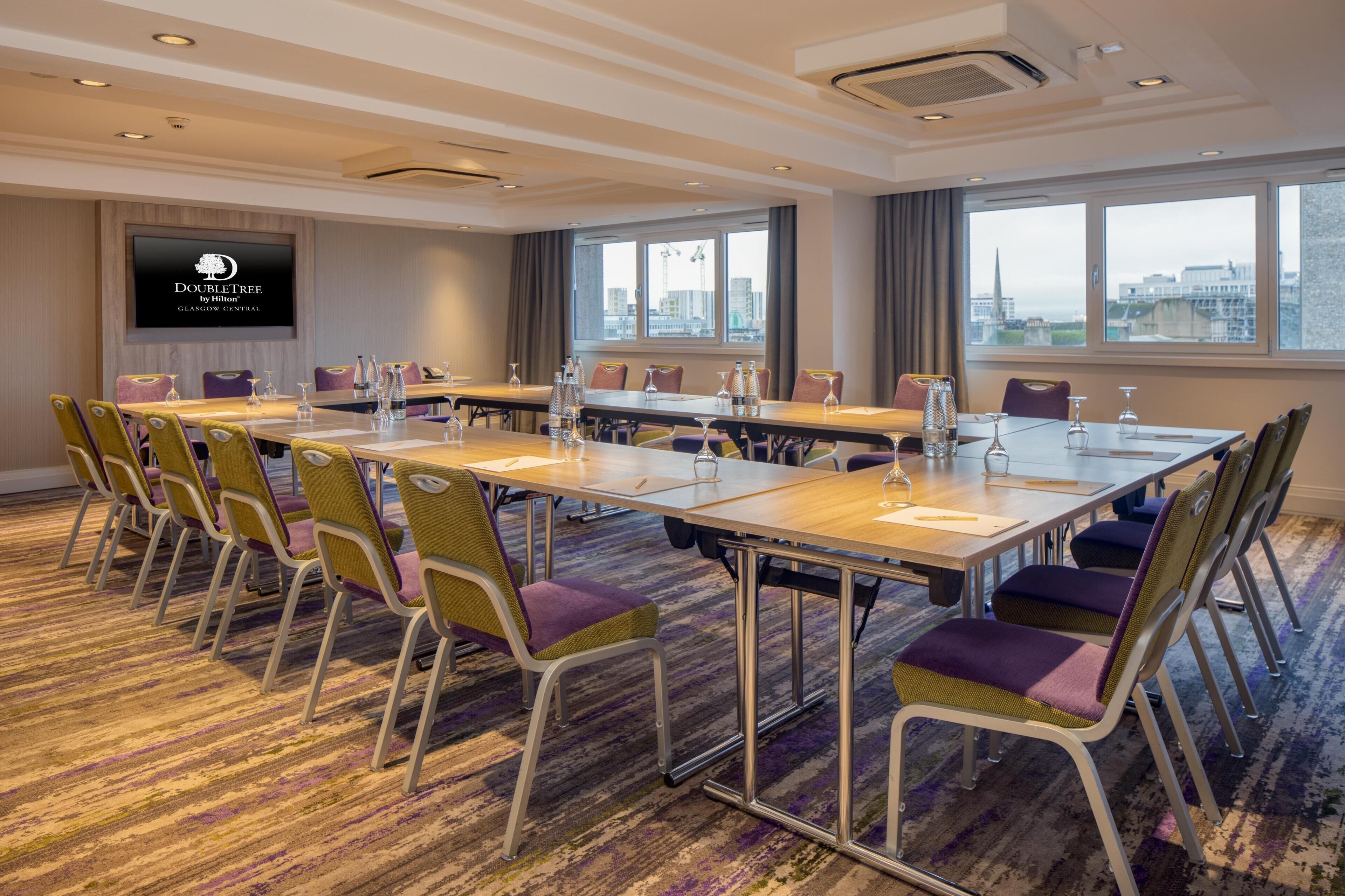 A conference room with a long table, multiple chairs, a flat-screen TV, and large windows with a city view.