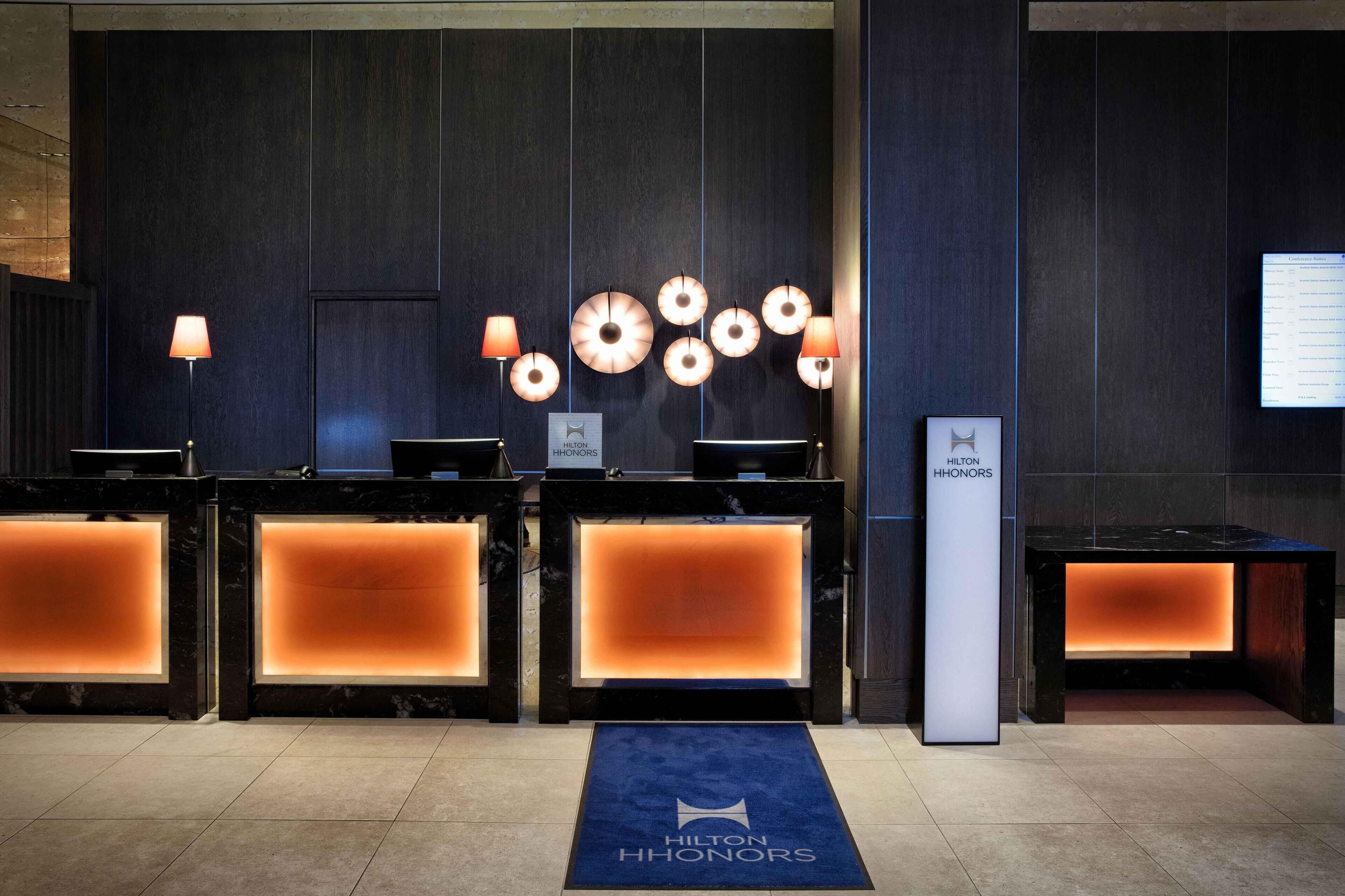 A hotel reception area with multiple reception desks, a blue carpet with the hotel name, and a wall-mounted display.