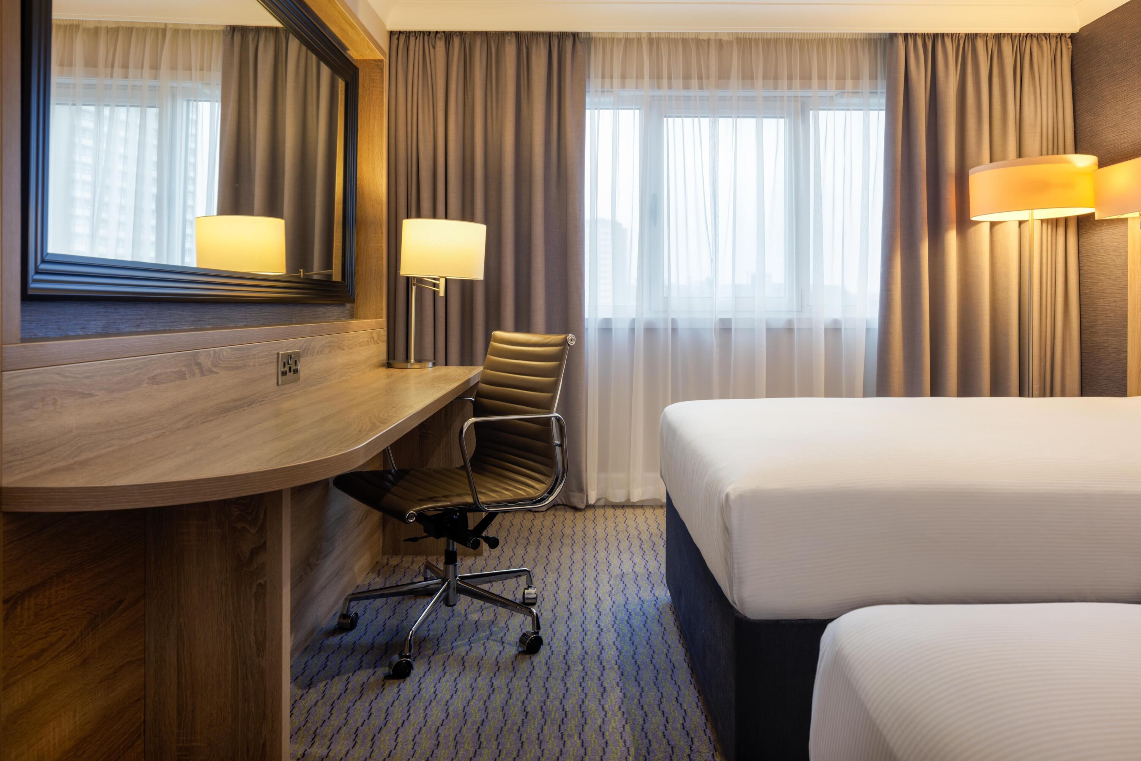 A hotel room with a wooden desk, a chair, a bed, a large mirror, and two lamps.