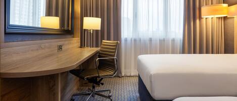 A hotel room with a wooden desk, a chair, a bed, a large mirror, and two lamps.