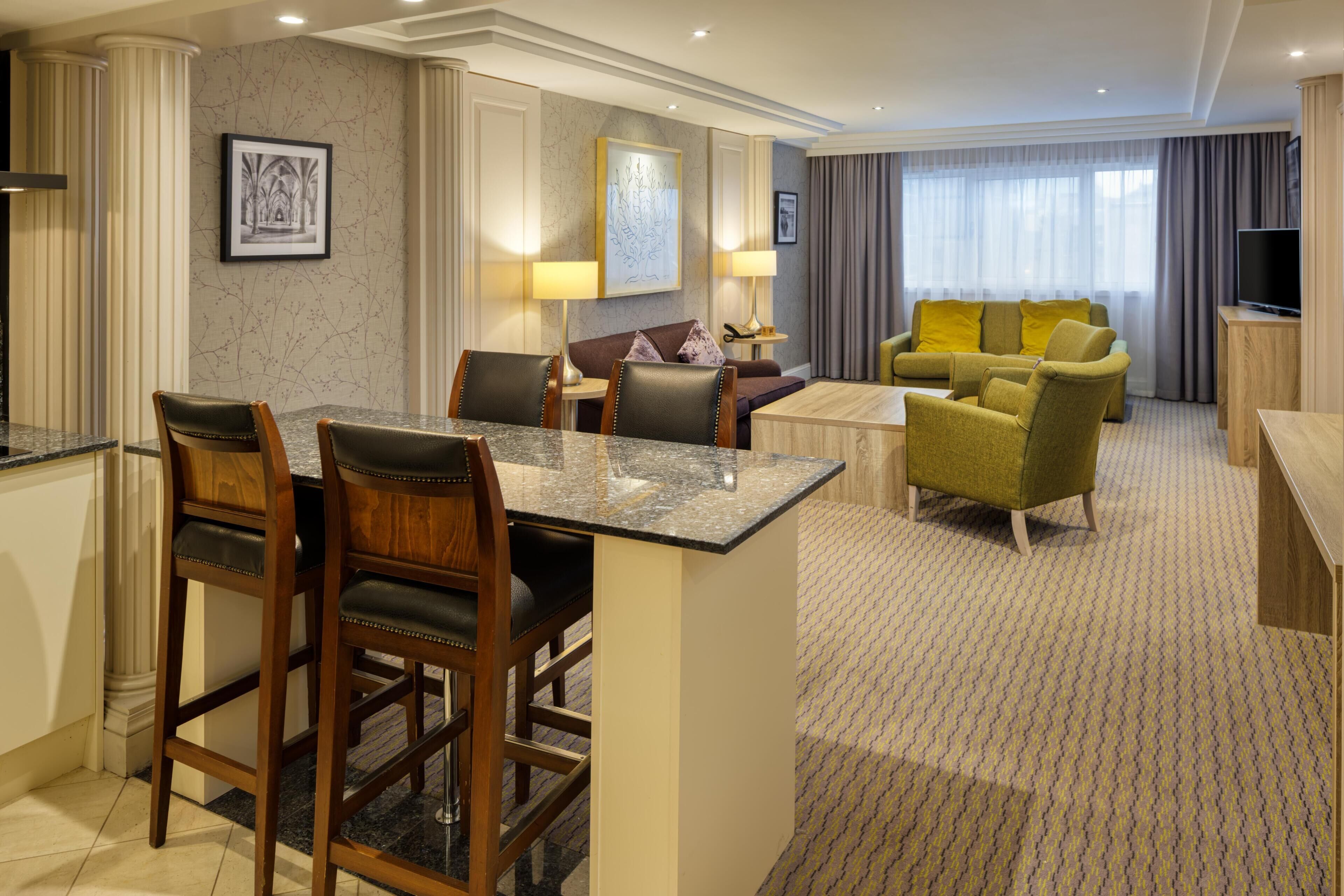 Habitación de hotel con zona de comedor, un sofá, televisor y un ventanal grande.