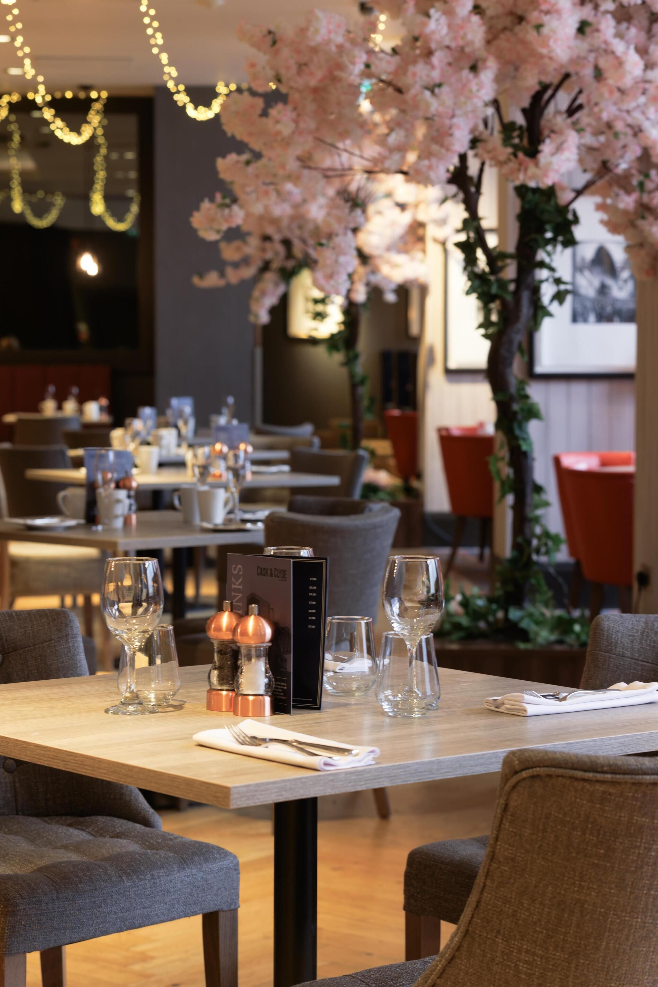 Un ambiente de restaurante con mesas dispuestas para comer, un atril para el menú y detalles decorativos como flores y luces colgantes.