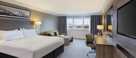 A hotel room with a large bed, a desk, a chair, a TV, and a view of the city.