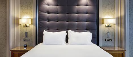 A hotel room with a large bed, two bedside lamps, a nightstand, and a wall-mounted alarm clock.