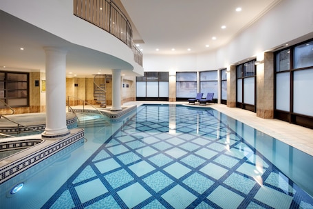 A large indoor swimming pool with a geometric tile pattern, a central staircase, and a balcony overlooking the pool.
