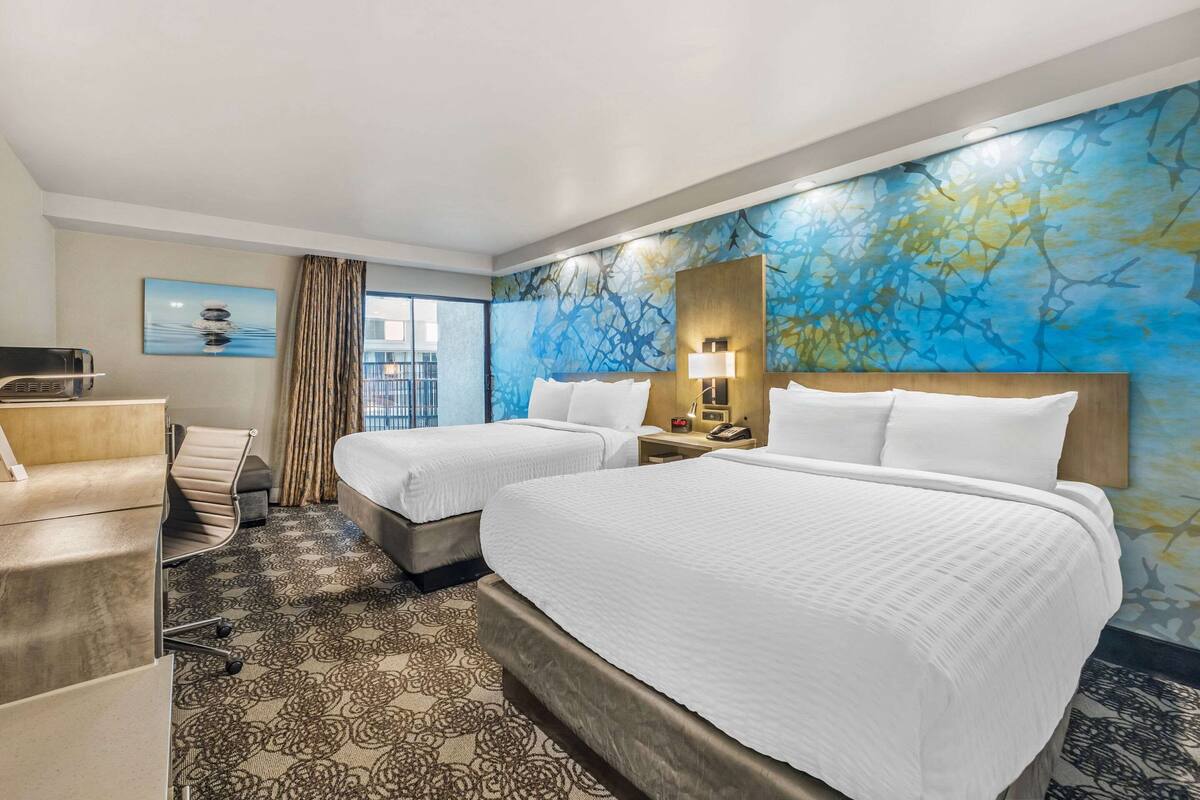 Une chambre d’hôtel avec deux lits, un bureau, une chaise, une grande fenêtre et un mur orné d’un motif floral bleu.