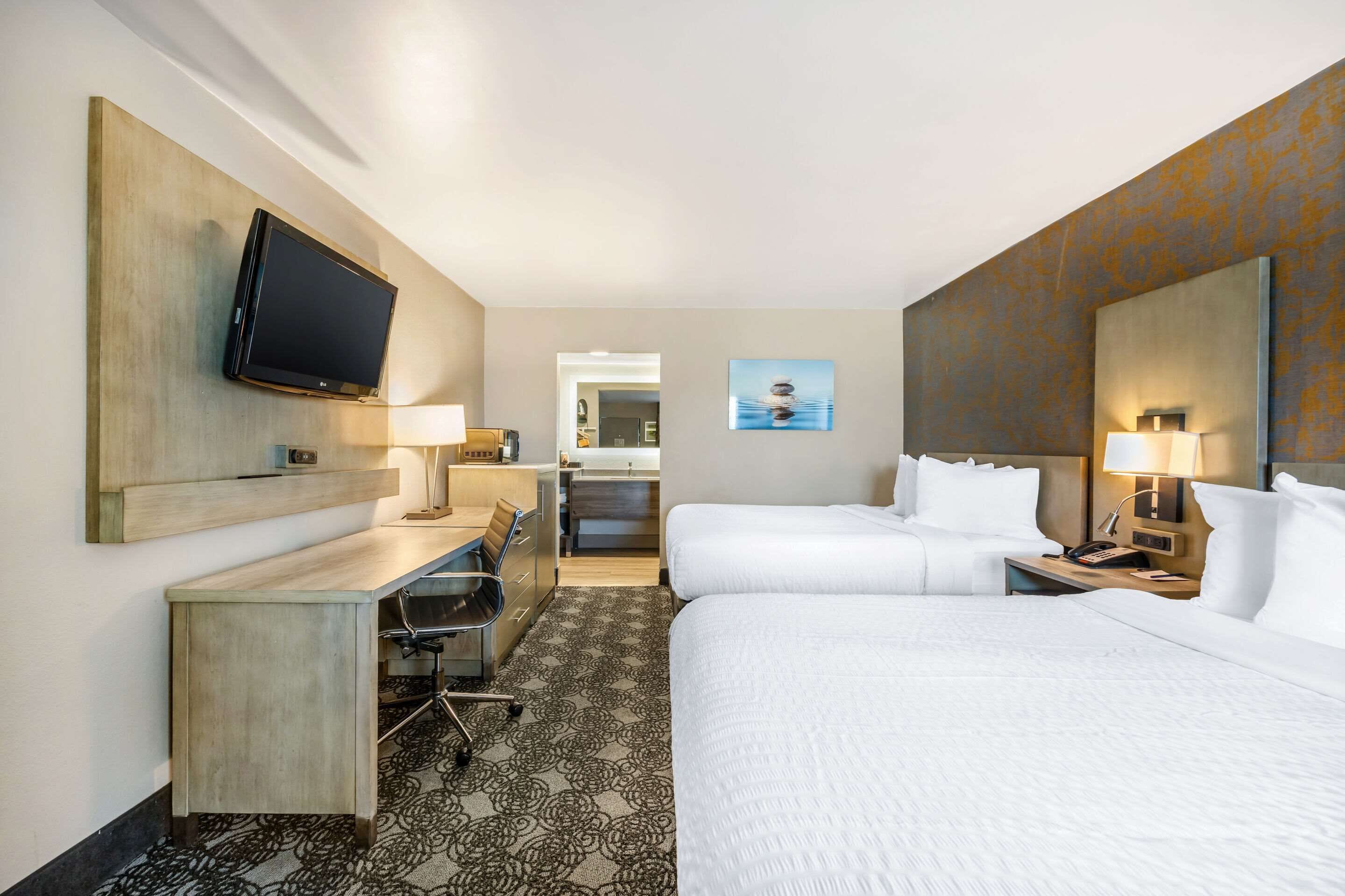 Hotelkamer met een flatscreen televisie aan de muur, een bureau met stoel, twee bedden met witte lakens en een kamerbreed tapijt met een patroon.