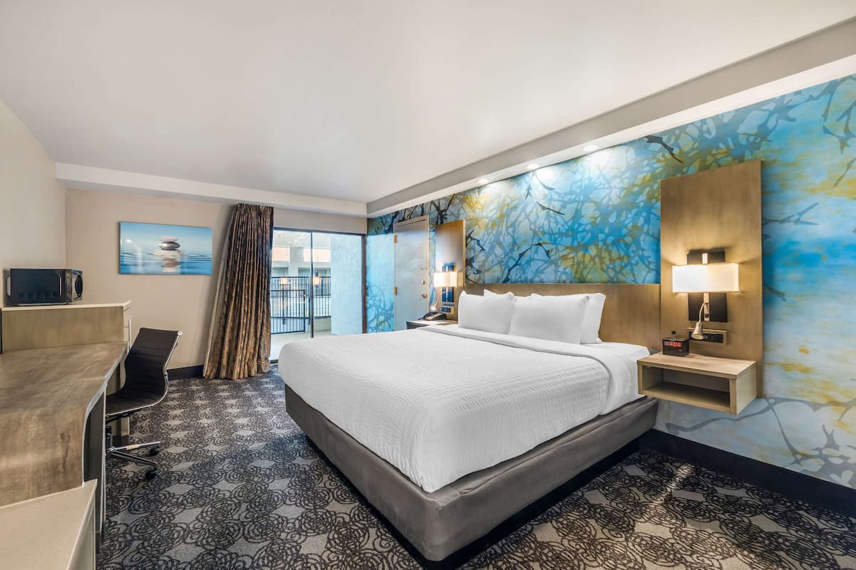 Une chambre d’hôtel avec un grand lit, un bureau et une chaise. Il y a une fenêtre avec des rideaux, un mur orné d’un motif abstrait bleu et une table de chevet avec une lampe.