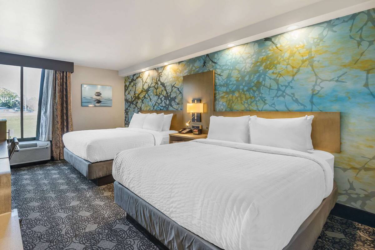 Une chambre d’hôtel avec deux lits, une grande fenêtre, un mur orné d’une fresque florale et une petite table avec une lampe.