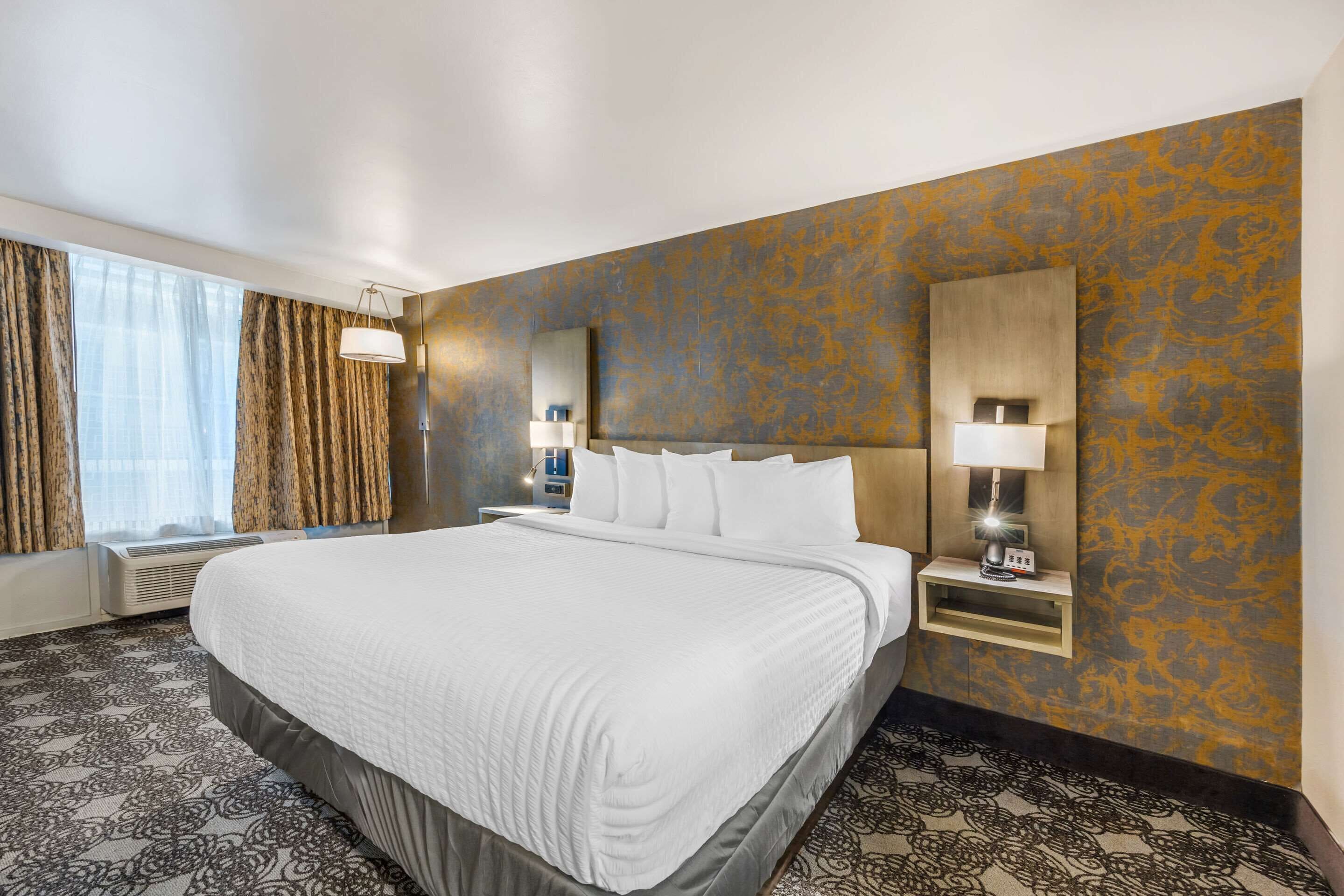 Habitación de hotel con una cama grande, una mesita de noche con lámpara, una ventana con cortinas y una pared con diseño decorativo.