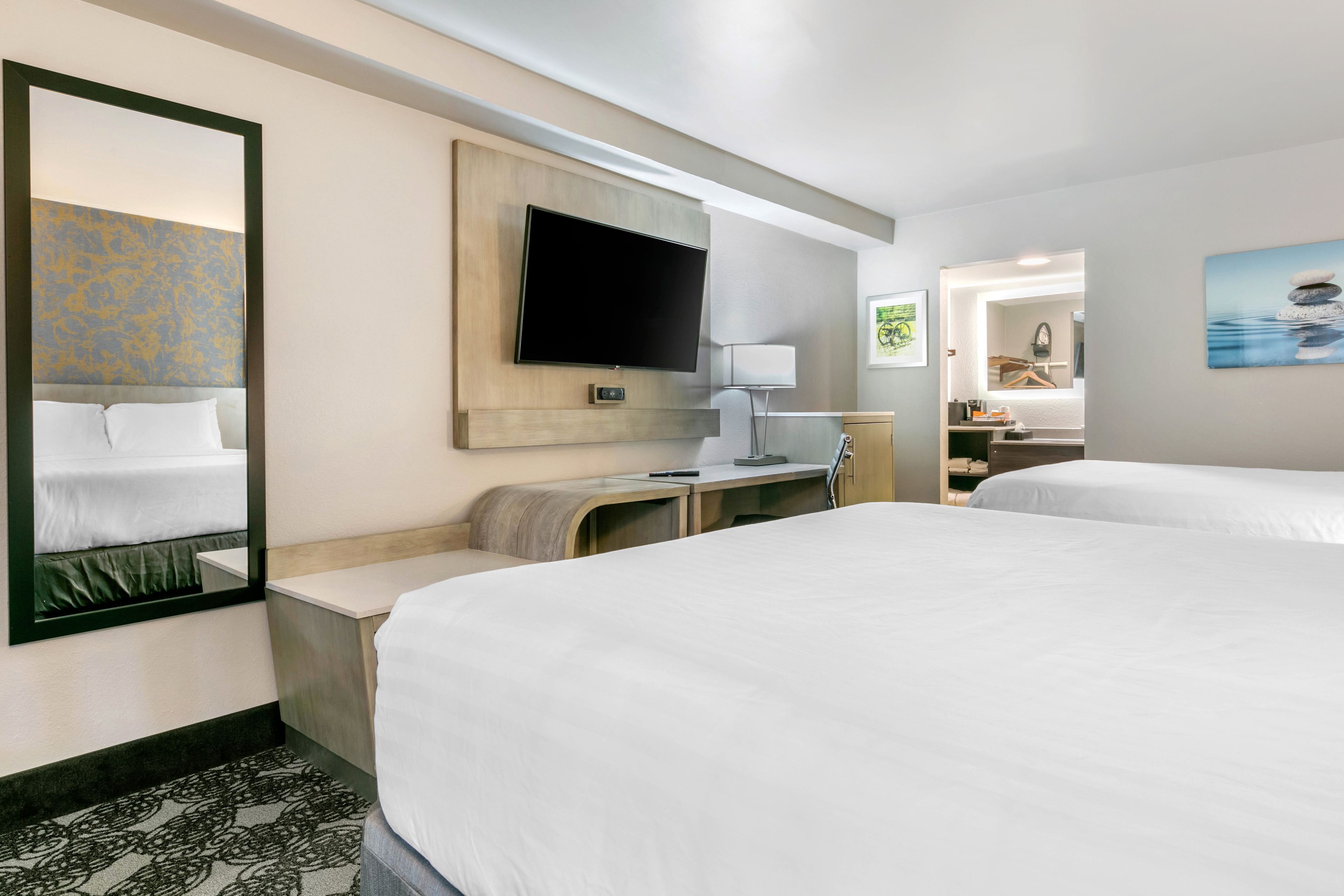 A hotel room with two beds, a TV mounted on the wall, a desk with a lamp, and a bathroom visible through an open door.