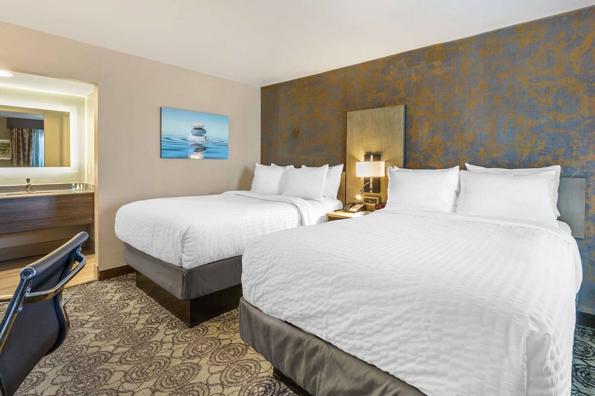 Une chambre d’hôtel avec deux lits, un coin salon avec un fauteuil, un grand miroir et un tableau au mur.
