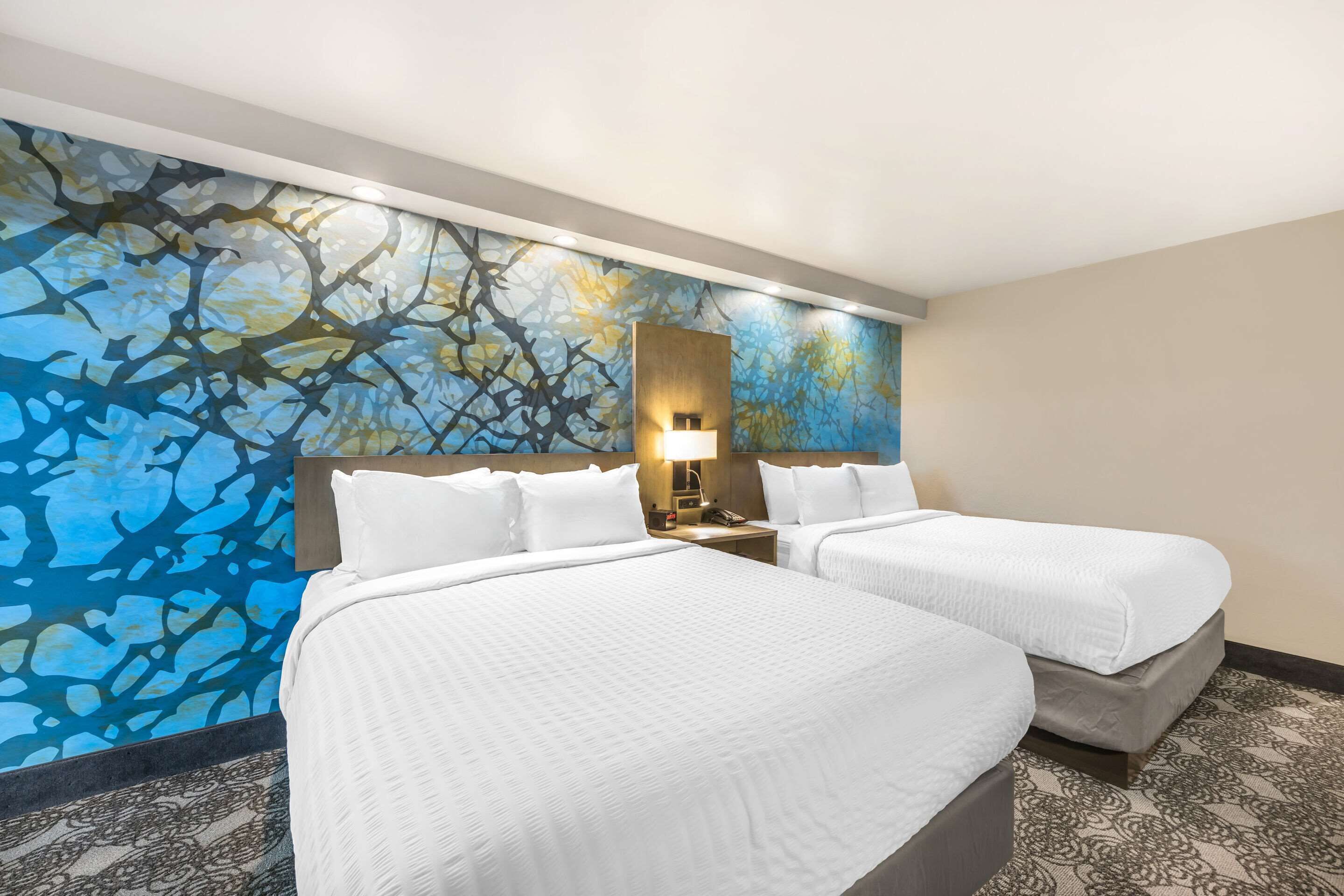 A hotel room with two beds, a large abstract mural on the wall, a nightstand with a lamp, and patterned carpet.