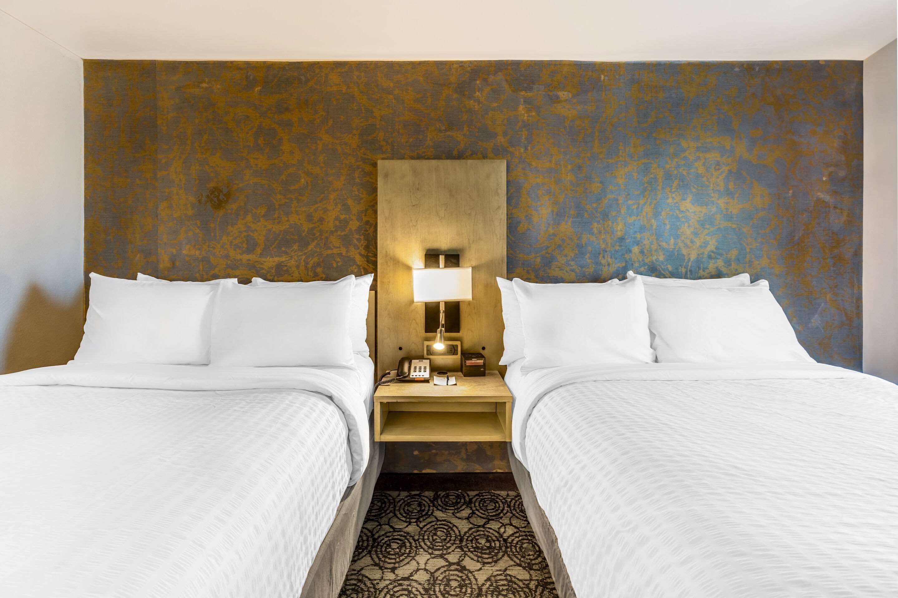 A hotel room with two beds, a nightstand with a lamp, and a textured wall.