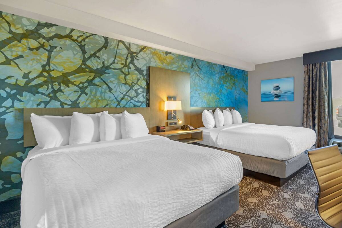 Une chambre d’hôtel avec deux lits, une grande fresque murale abstraite, un tableau représentant un paysage aquatique et une vue sur l’extérieur.