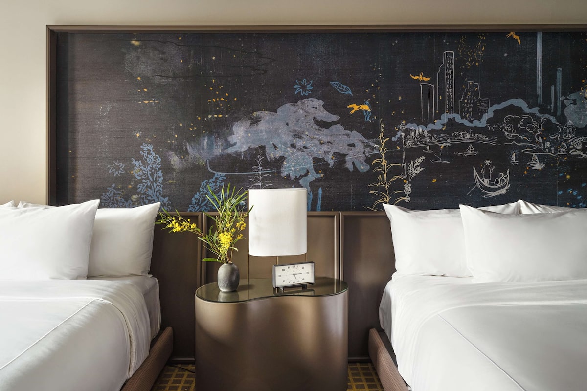 A hotel room with two beds, a nightstand with a lamp, and a large abstract mural on the wall.