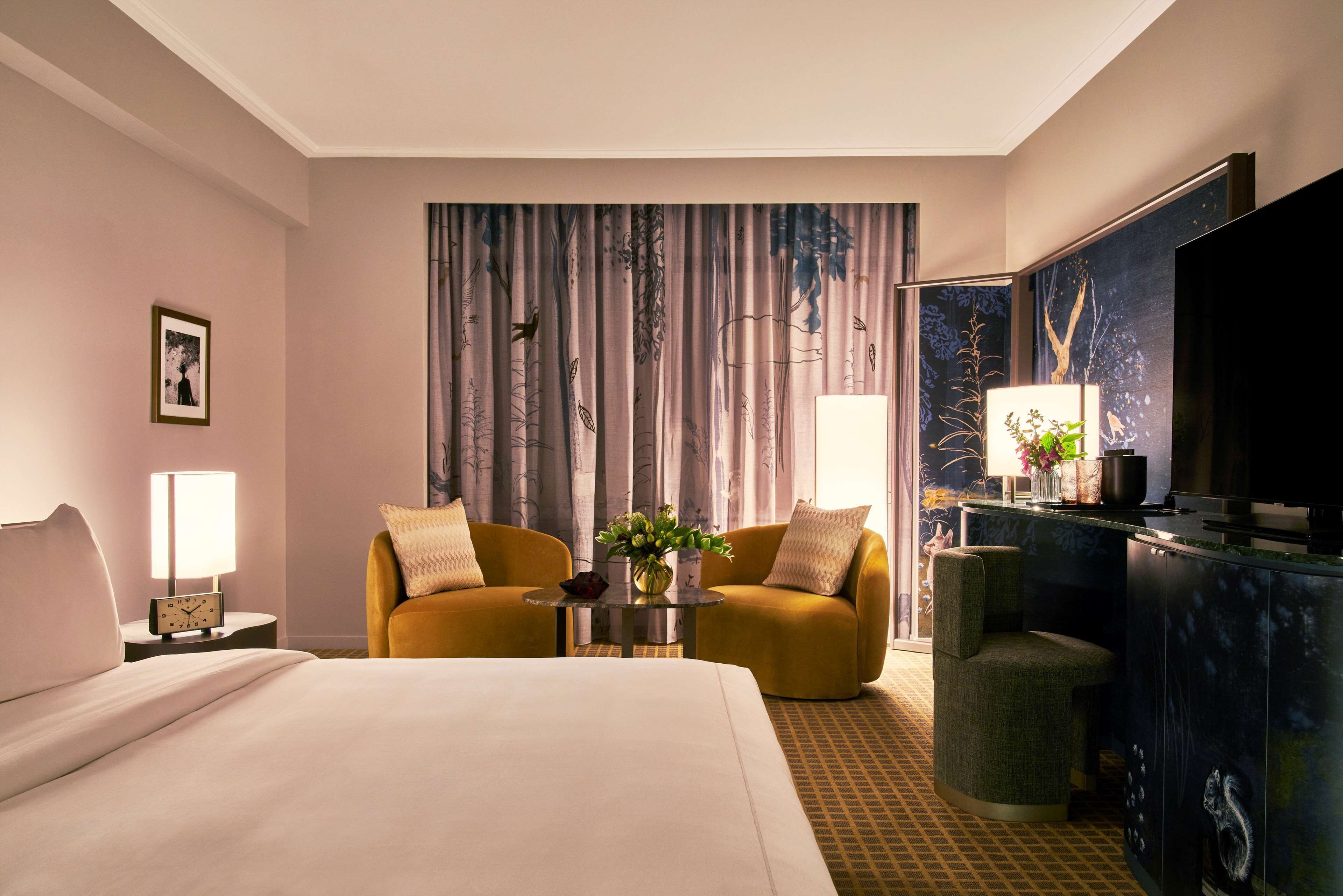 A hotel room with a bed, two armchairs, a small table with a vase of flowers, a lamp, a television, and a large window with curtains.