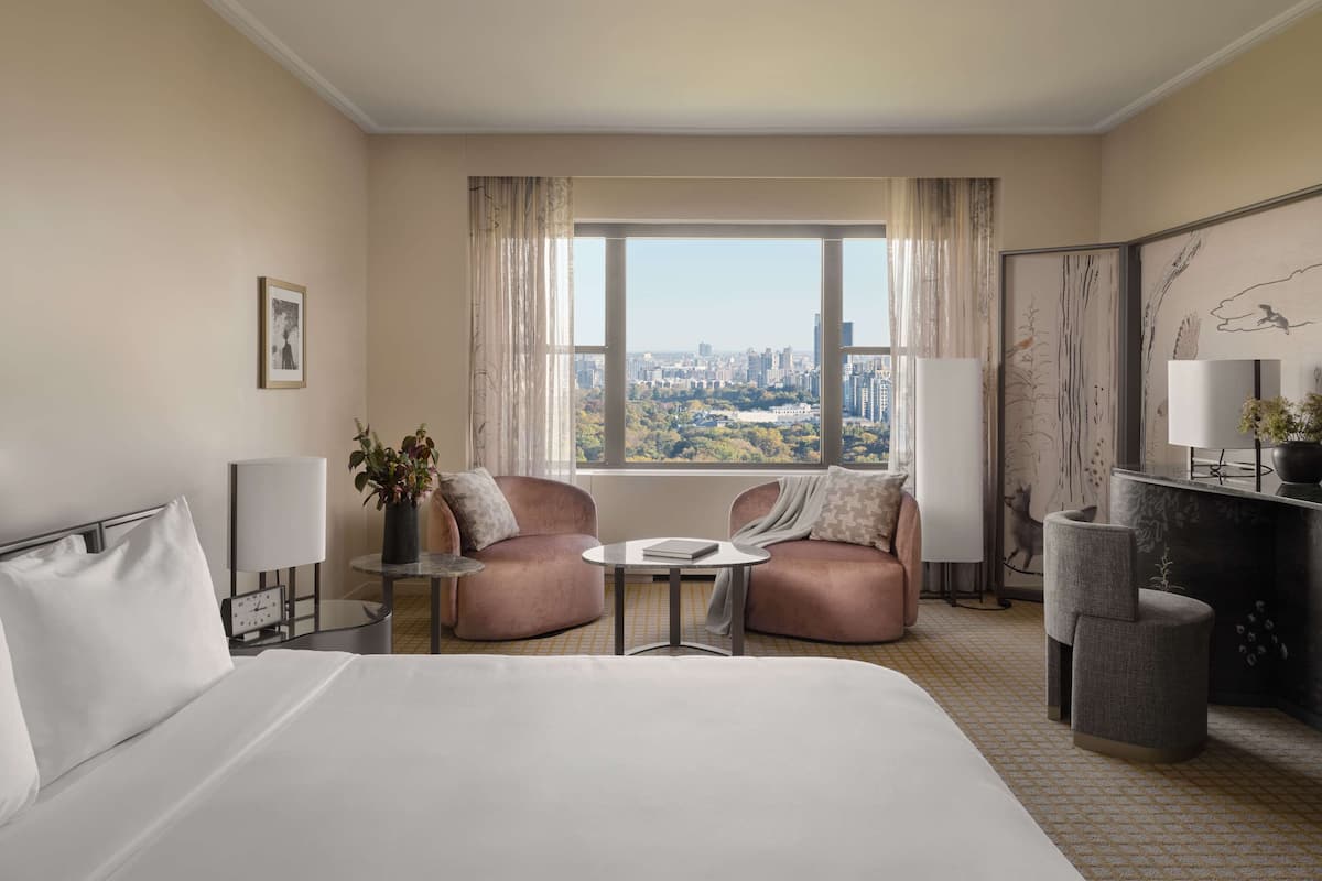 A hotel room with a bed, two armchairs, a small table, a desk with a lamp, and a view of a cityscape through large windows.
