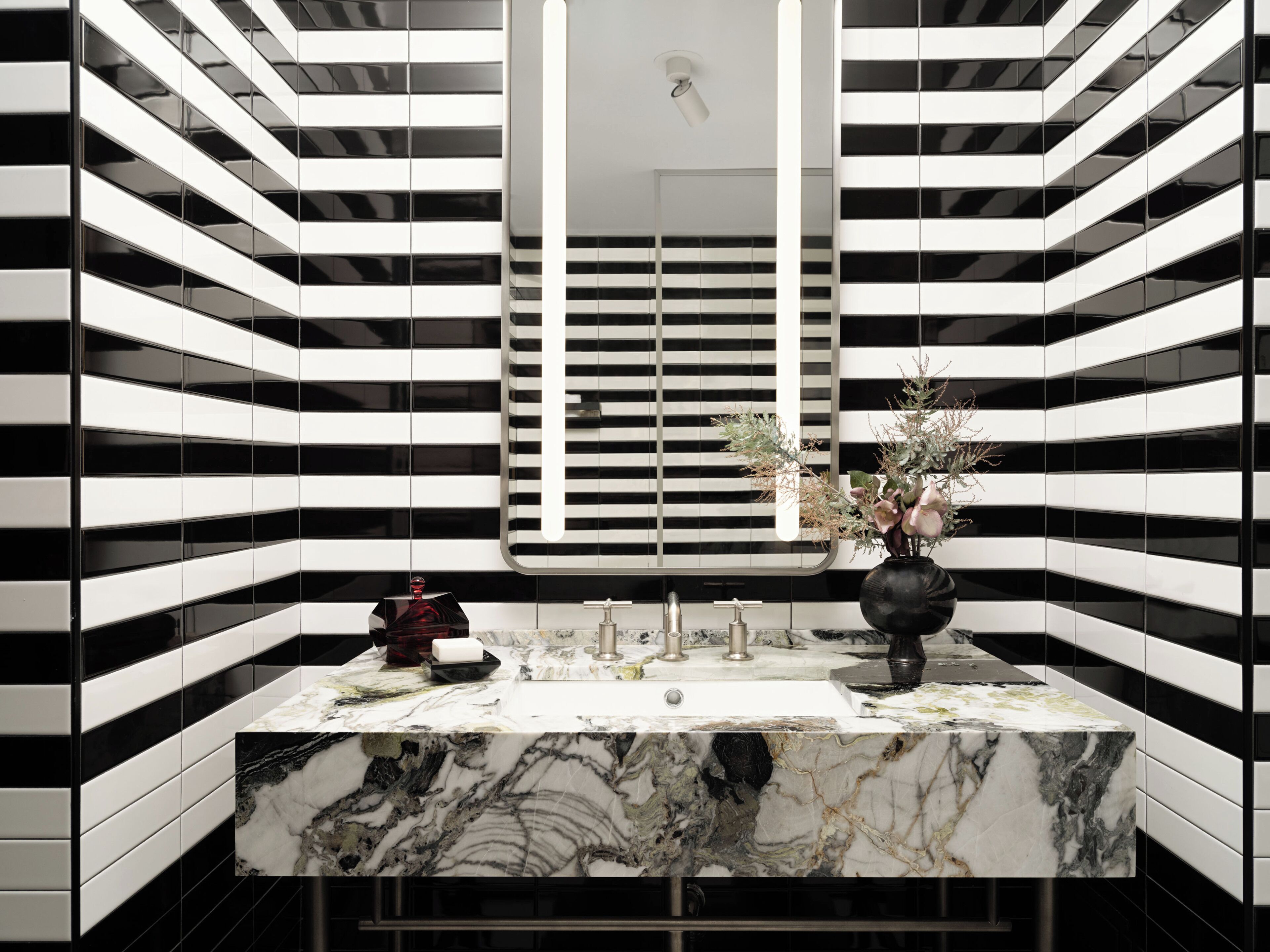 Un baño moderno con diseño de pared a rayas negras y blancas, un lavamanos de mármol y un florero con flores.