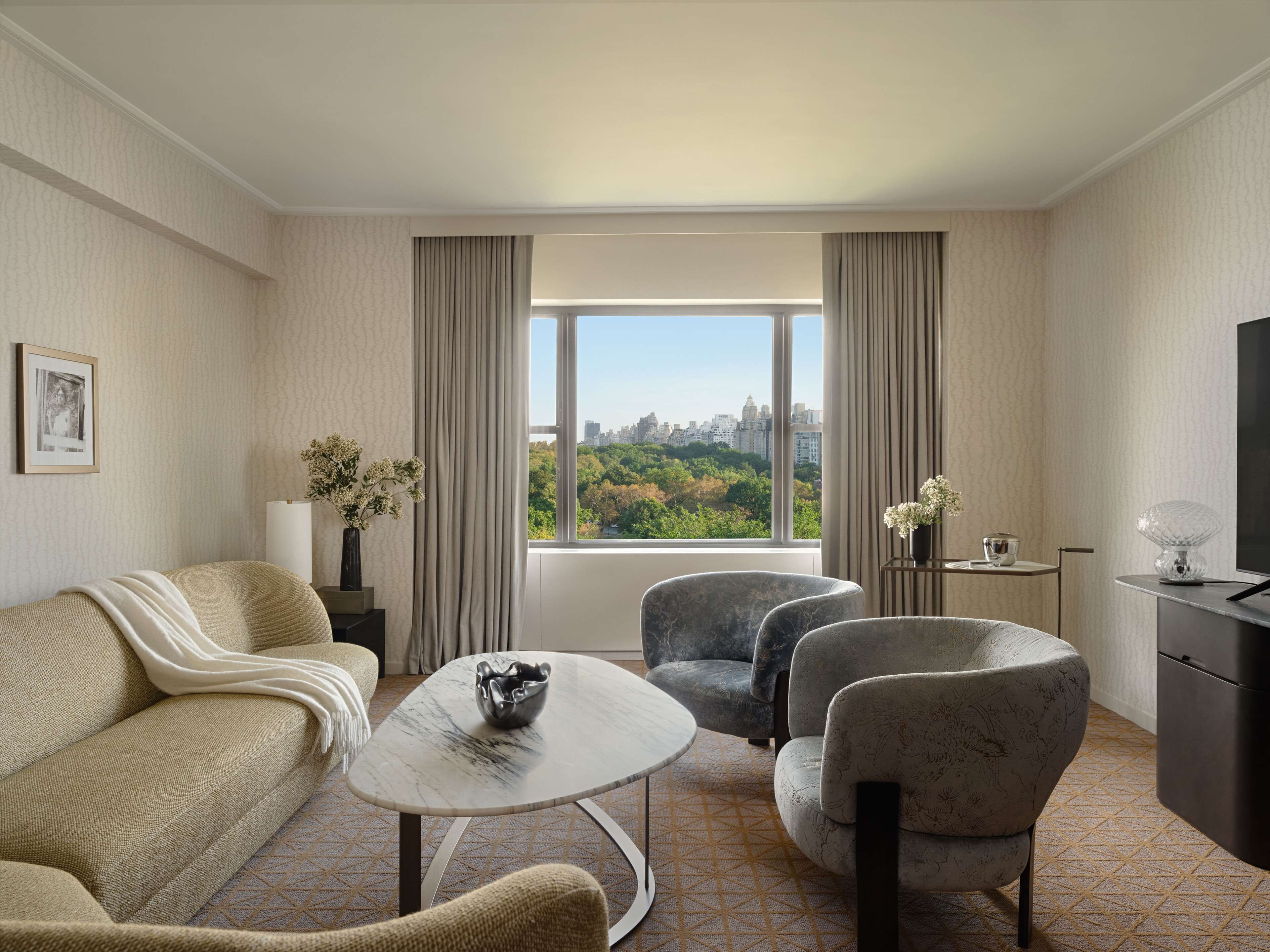 Une chambre d’hôtel moderne équipée d’un canapé, de deux fauteuils, d’une petite table, d’un téléviseur et d’une grande fenêtre donnant sur la ville.