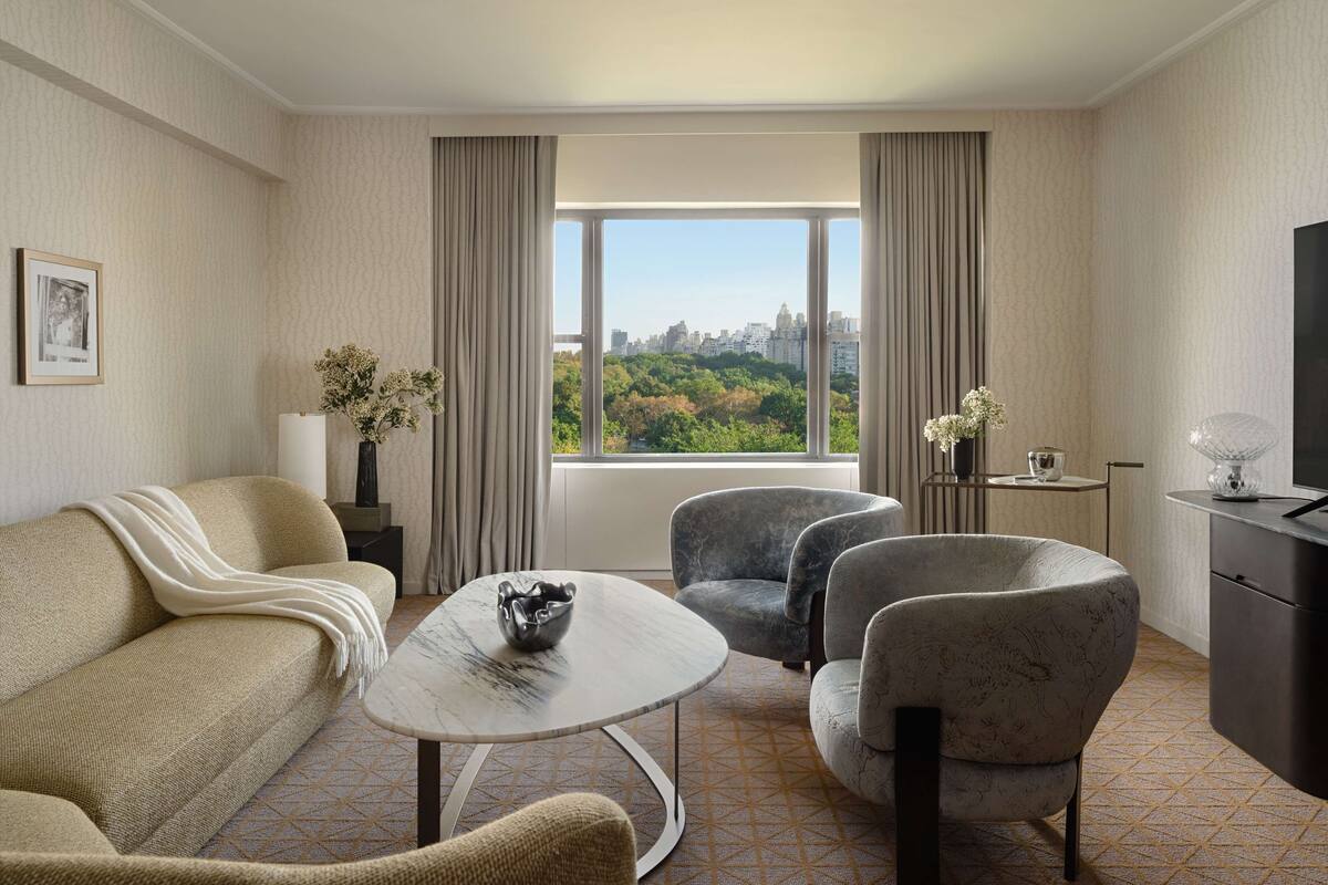 A modern hotel room with a sofa, two armchairs, a small table, a TV, and a large window with a city view.