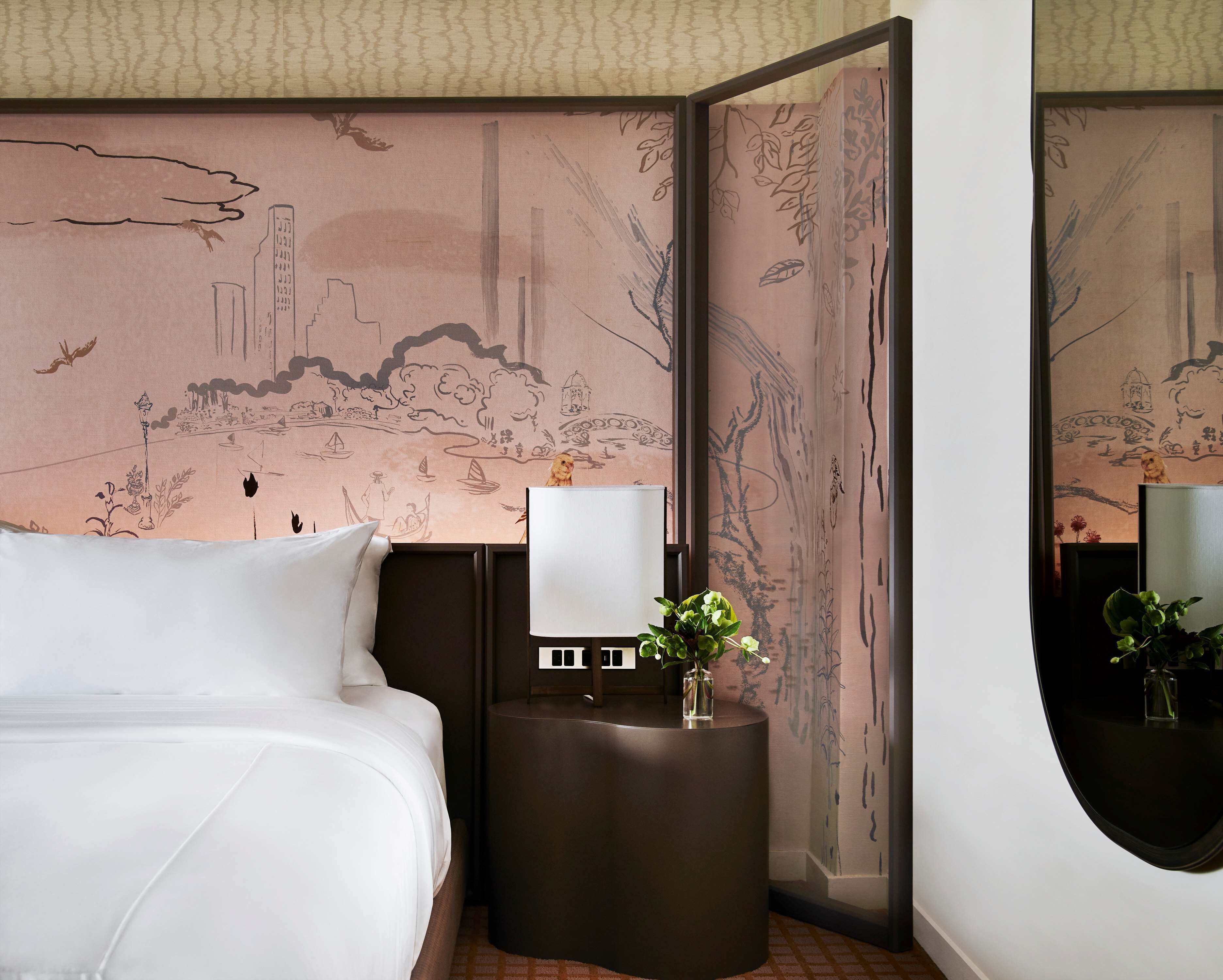 Ein Hotelzimmer mit einem Bett, einem Nachttisch mit Lampe, einer Vase mit Blumen und einer Wand mit Wandgemälde.