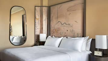 A neatly made bed with white linens, a round mirror, a wall-mounted artwork, and a bedside table with a lamp and a clock.