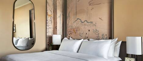 Una cama bien hecha con sábanas blancas, un espejo redondo, una obra de arte en la pared y una mesita de noche con una lámpara y un reloj.