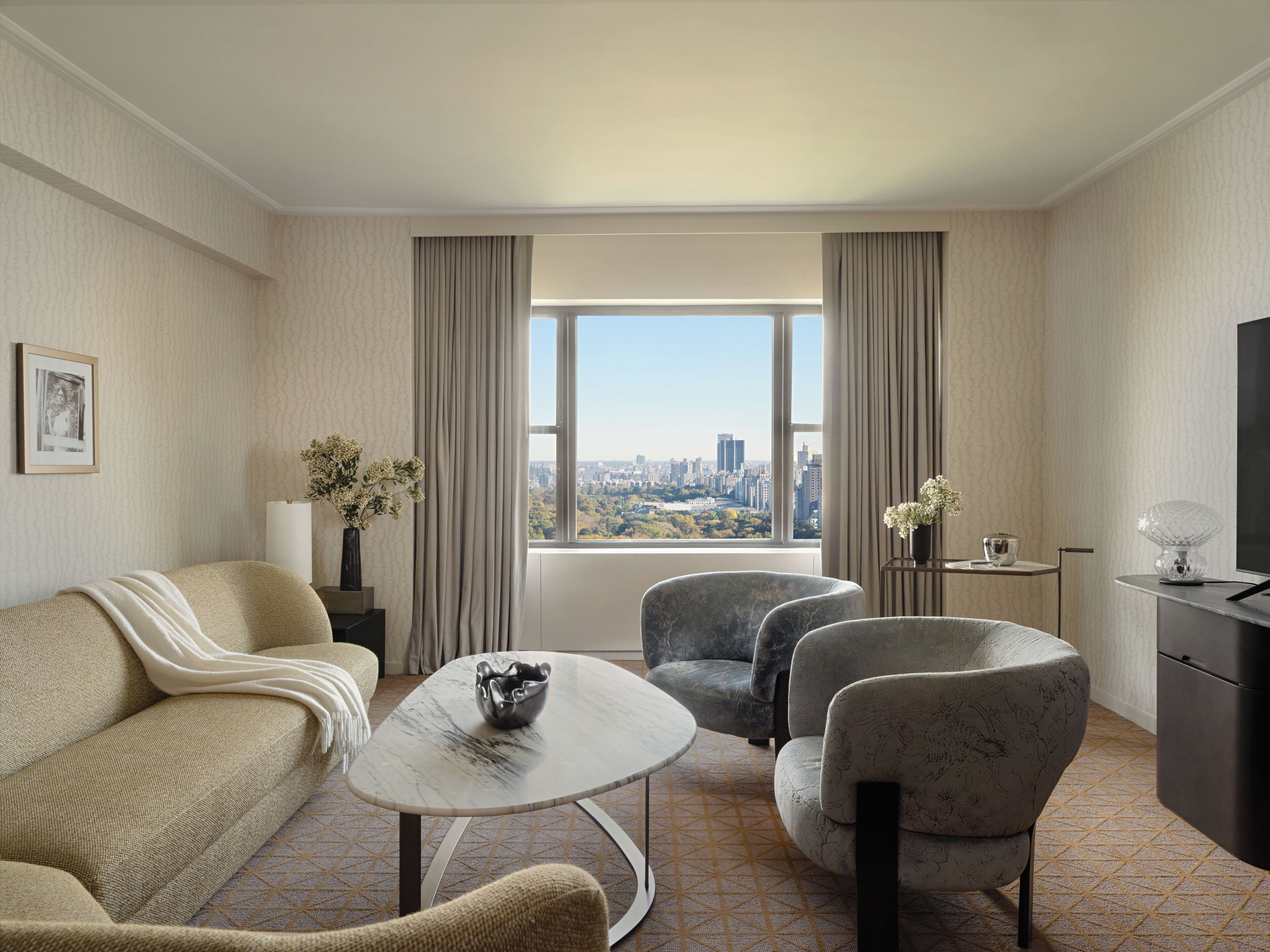 A hotel room with a sofa, two armchairs, a coffee table, a TV, and a large window with a city view.