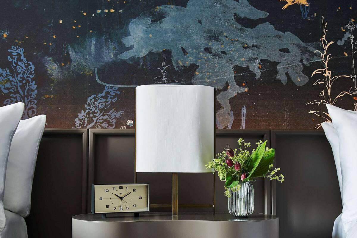 A modern hotel room with a dark brown console table, a white lamp, a clock, and a vase with flowers.