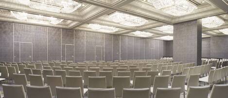 Une salle de conférence avec des rangées de chaises, un plafond haut orné de lustres et une décoration aux couleurs neutres.