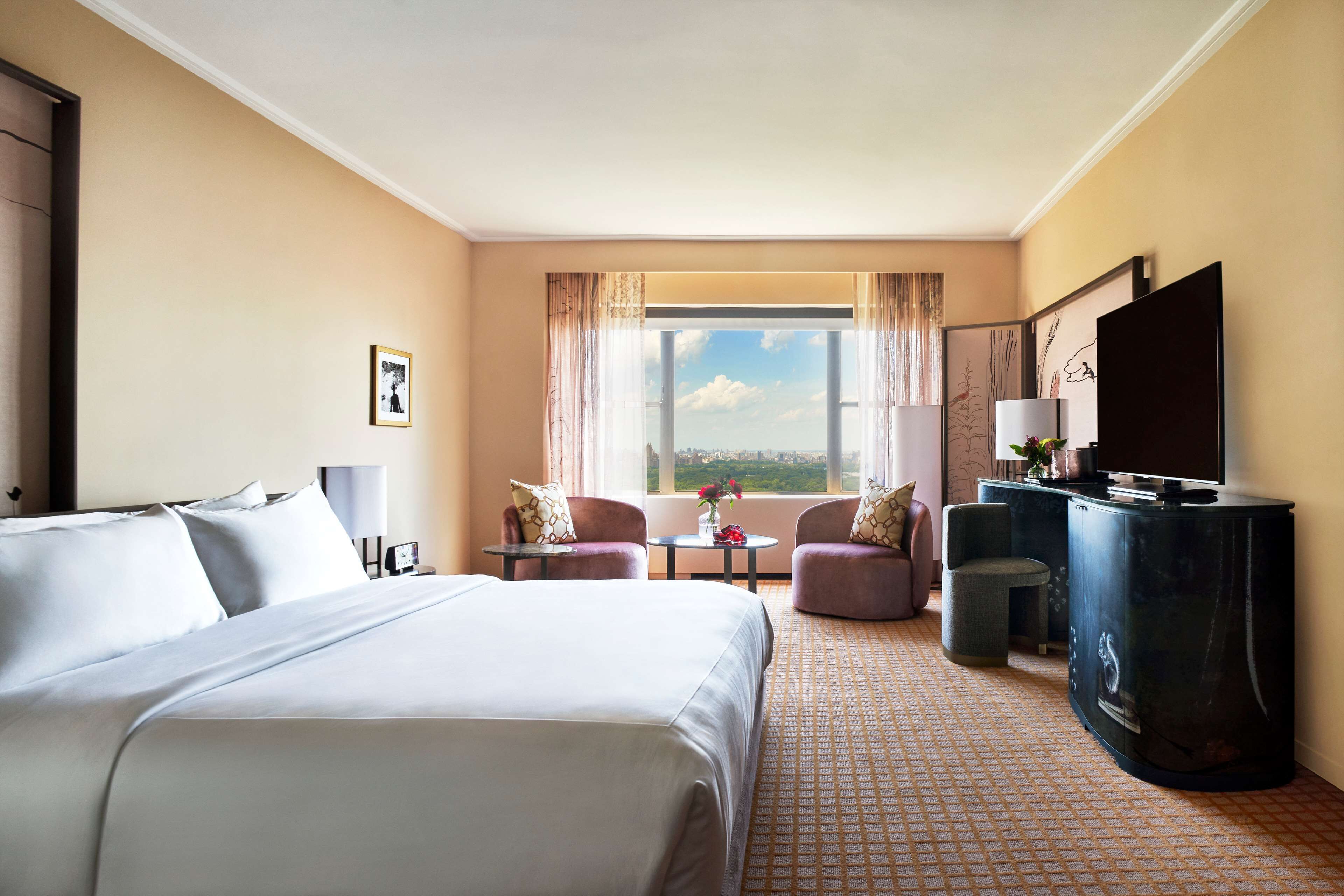 A hotel room with a large bed, two armchairs, a small round table, a TV, and a view of the city.