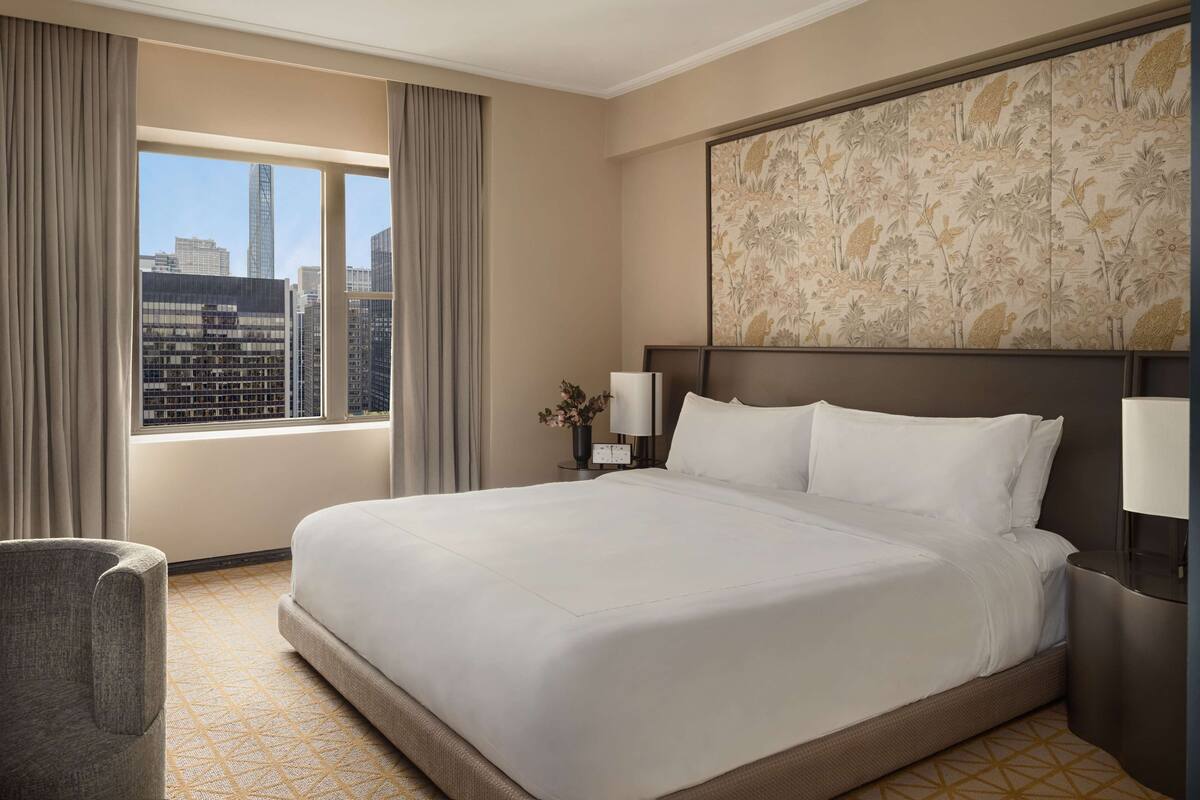 A hotel room with a large bed, a nightstand, a lamp, a chair, a window with curtains, and a city view.