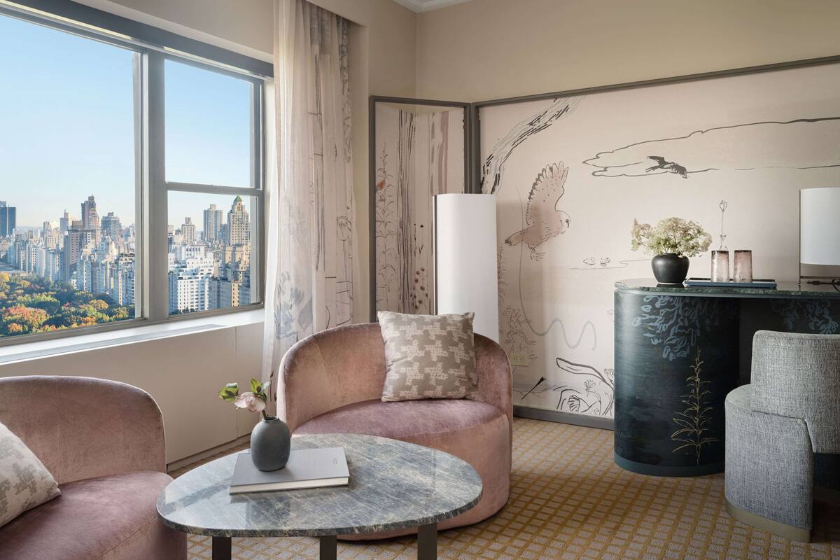 A modern hotel room with a large window offering a city view, a comfortable seating area with a round table, and a decorative wall with an owl mural.