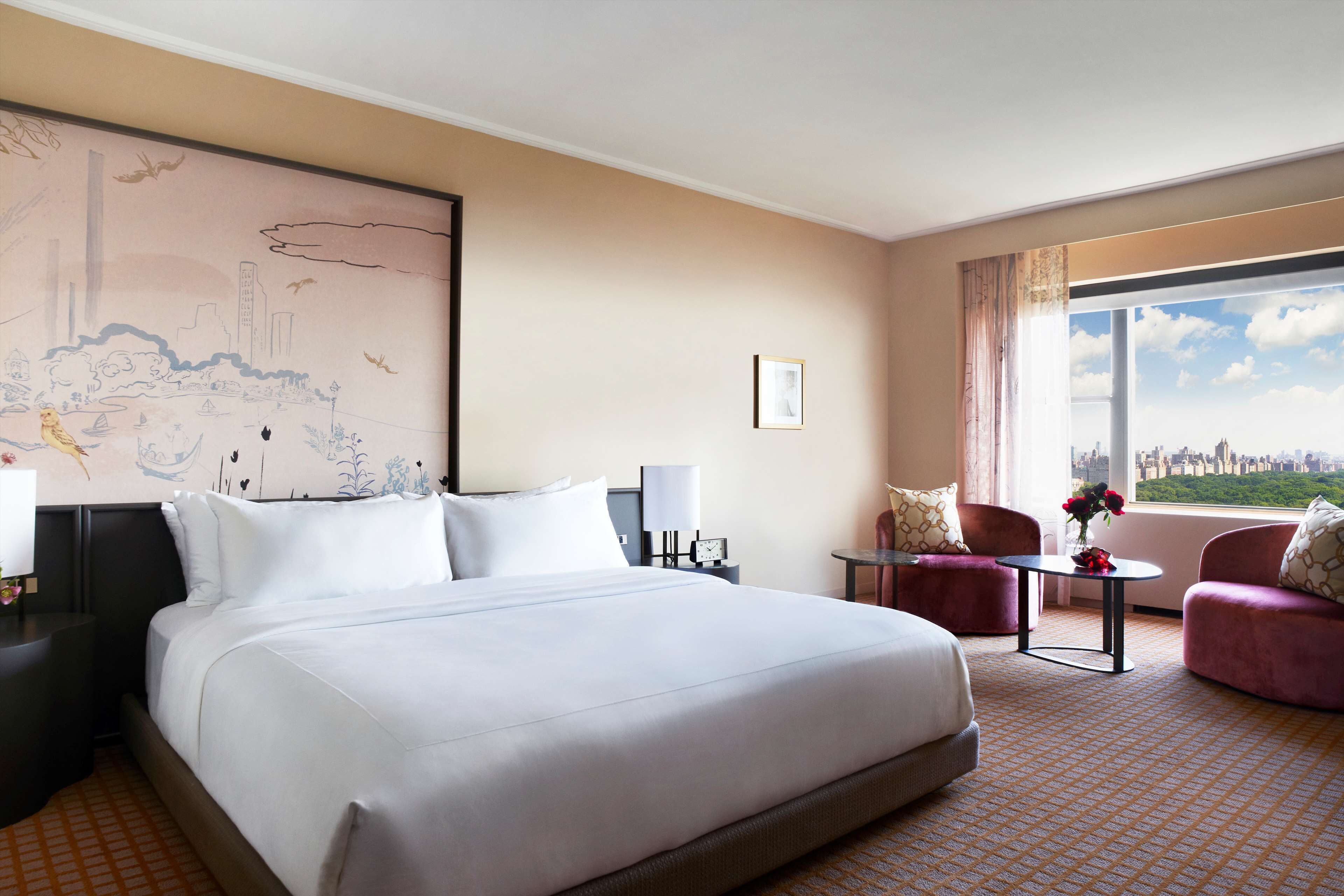 A hotel room with a large bed, two armchairs, a small table with flowers, a cityscape view, and a large wall mural.
