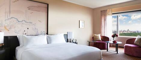Habitación de hotel con una cama grande, dos sillones, una mesita con flores, vistas a la ciudad y un mural grande en la pared.