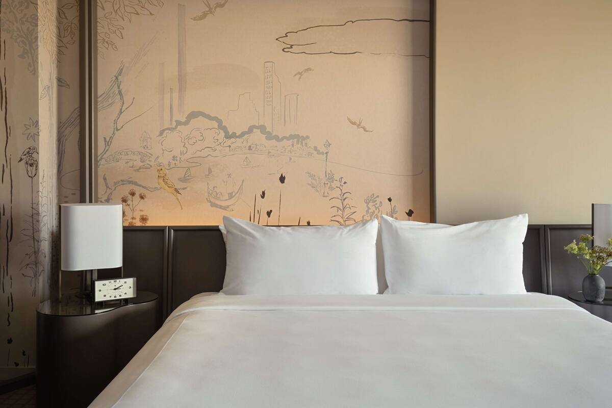 A hotel room with a large bed, a nightstand with a lamp, and a wall mural depicting a landscape with birds and boats.