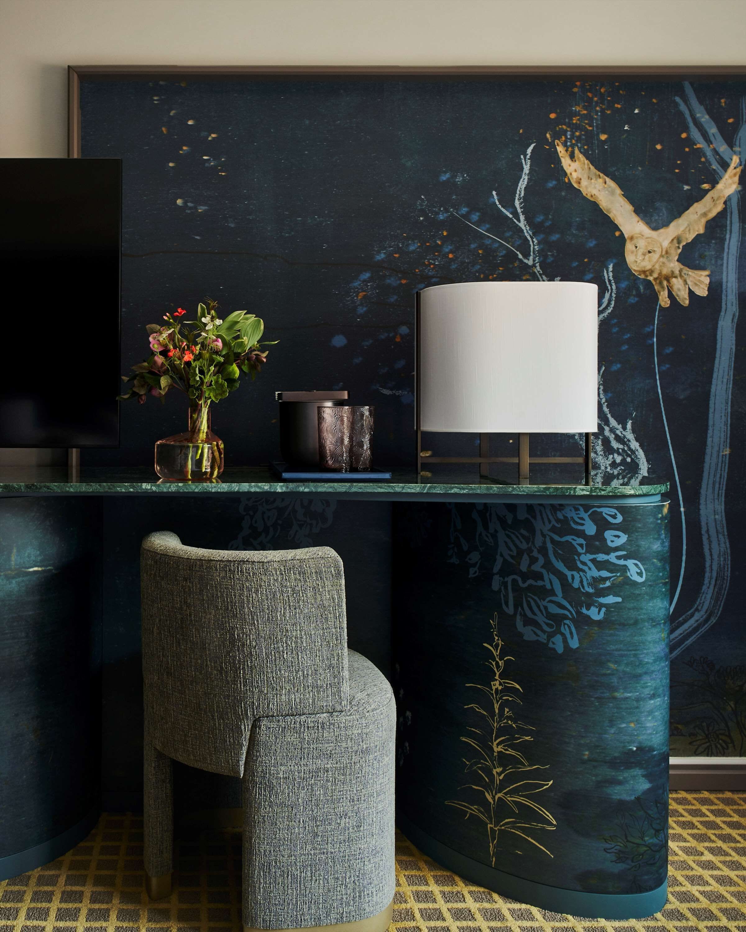 Una habitación moderna con un mural de tonos oscuros, un televisor, un florero con flores, una lámpara y una silla.