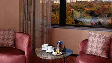 Een hotelkamer met twee comfortabele fauteuils, een klein rond tafeltje met een koffiezetapparaat, kopjes en een bord eten, en een groot raam met uitzicht op een stads panorama en herfstkleuren.